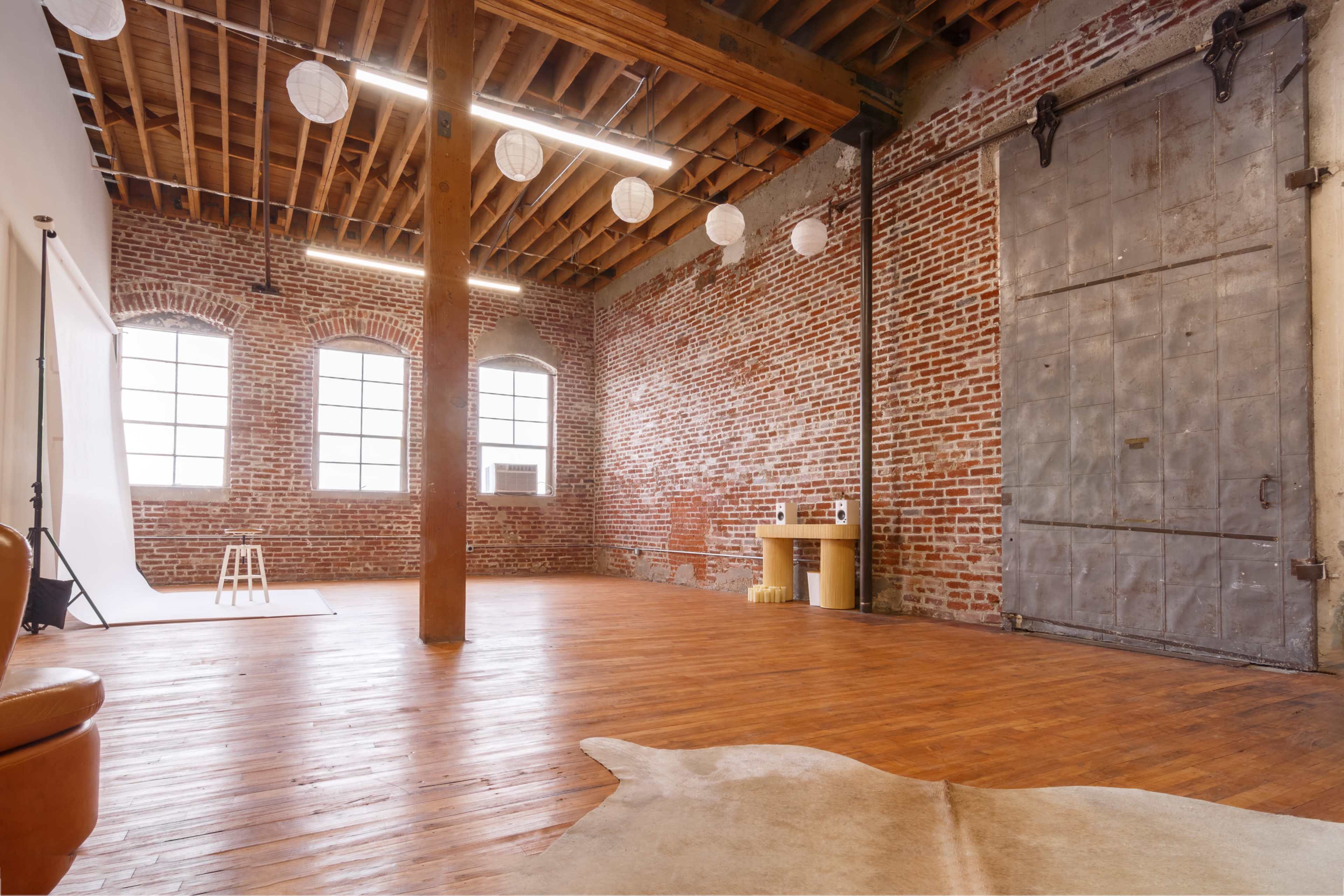 A spacious room with exposed brick walls, wooden beams, and large windows, featuring a wooden floor and a simple arrangement of furniture.