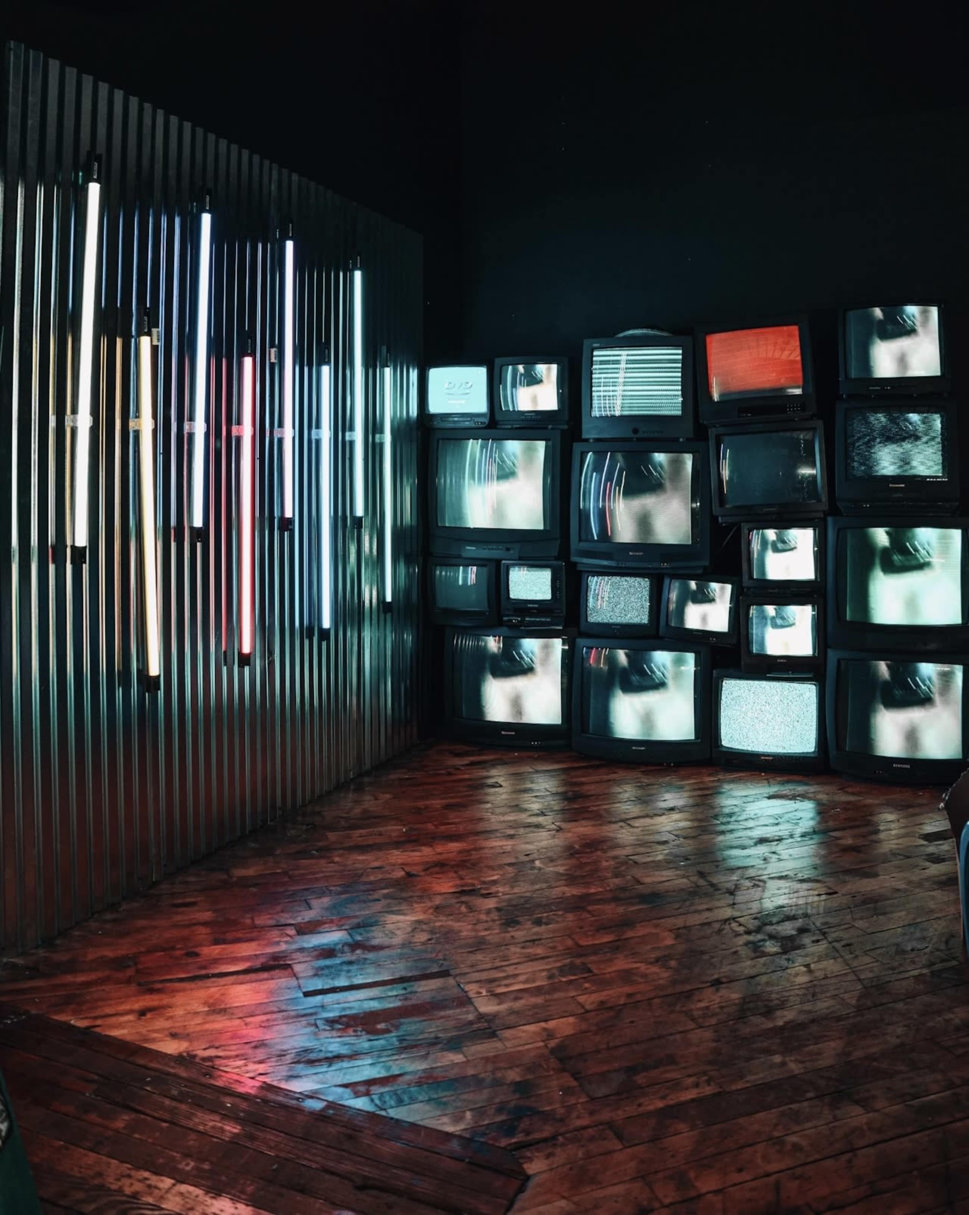 A corner of a room features multiple vintage televisions displaying various static images, alongside a wall of illuminated neon lights.