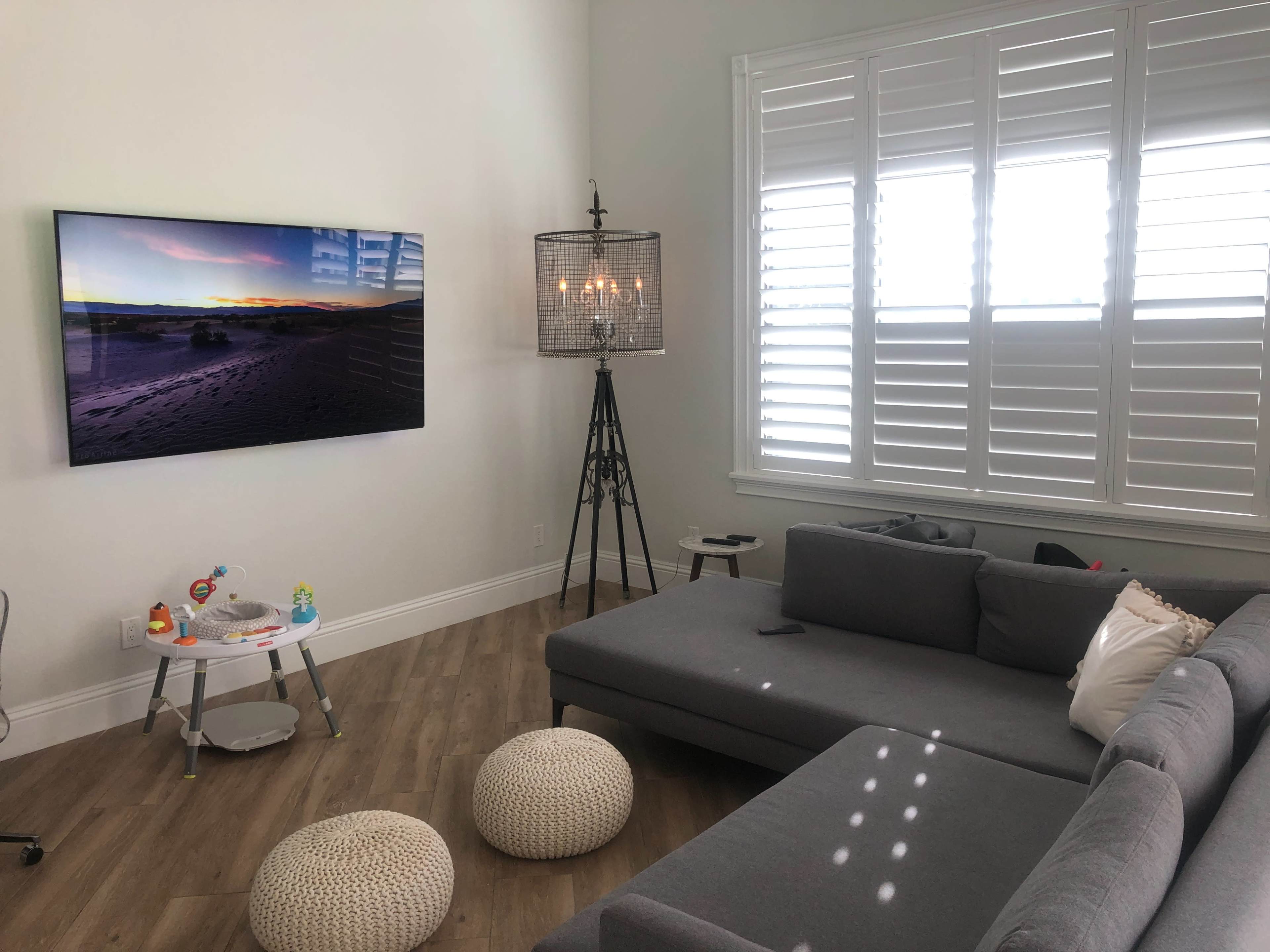 A modern living room features a large TV mounted on the wall, a gray sectional sofa, a side table with toys, and a standing lamp beside a window with white shutters.