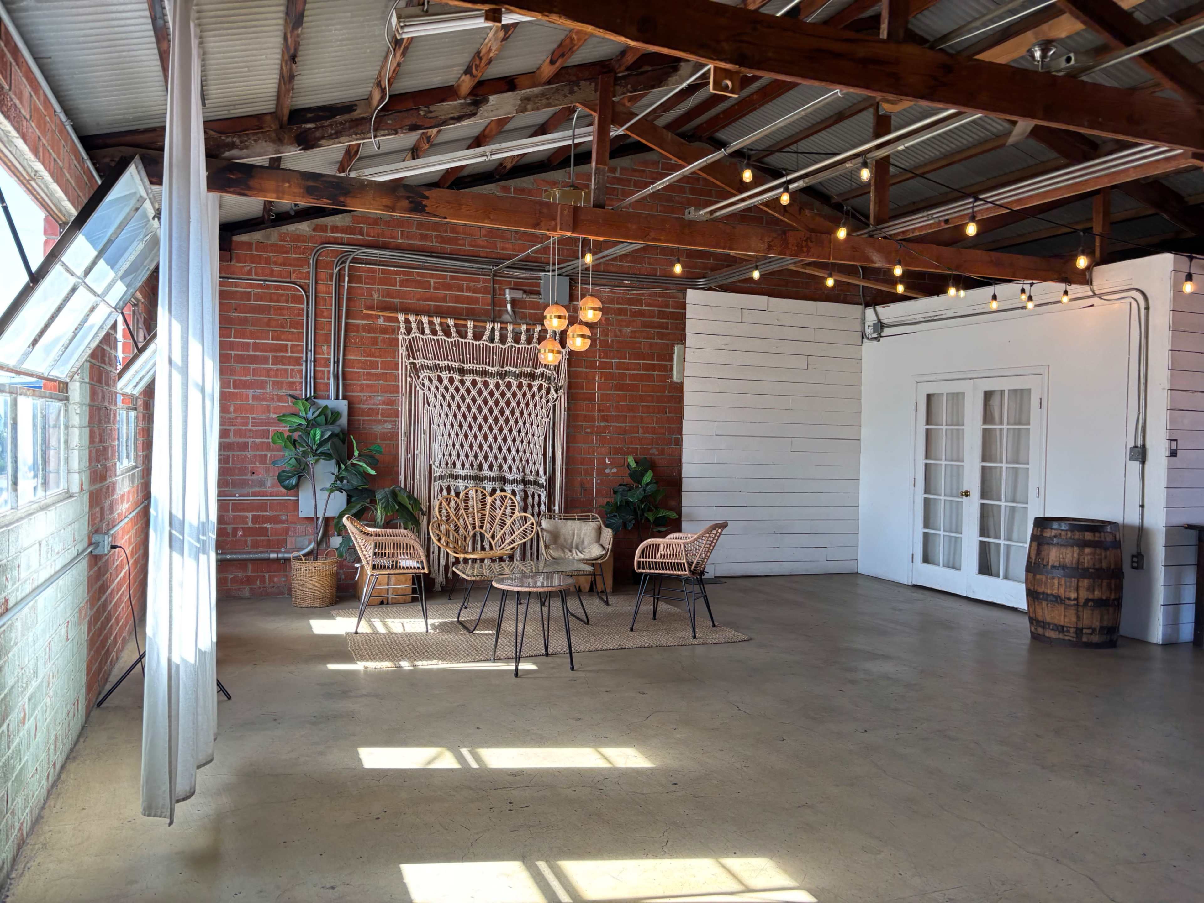 The image shows a spacious, stylish interior with exposed brick walls, wooden beams, and light fixtures, furnished with wicker chairs, a round table, and decorative plants.