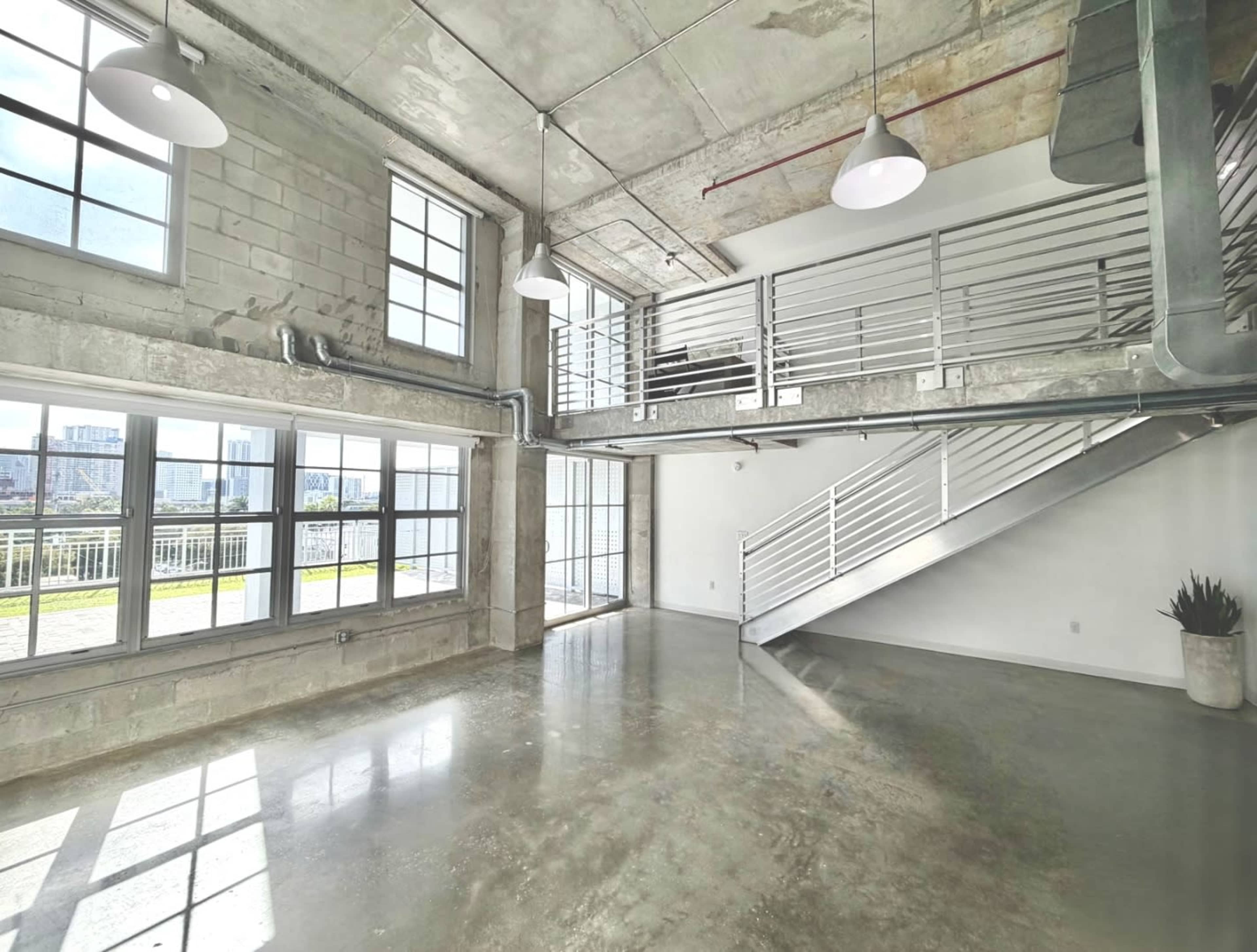 The image shows an open industrial-style space with concrete walls, large windows, and a stairs leading to a mezzanine level.