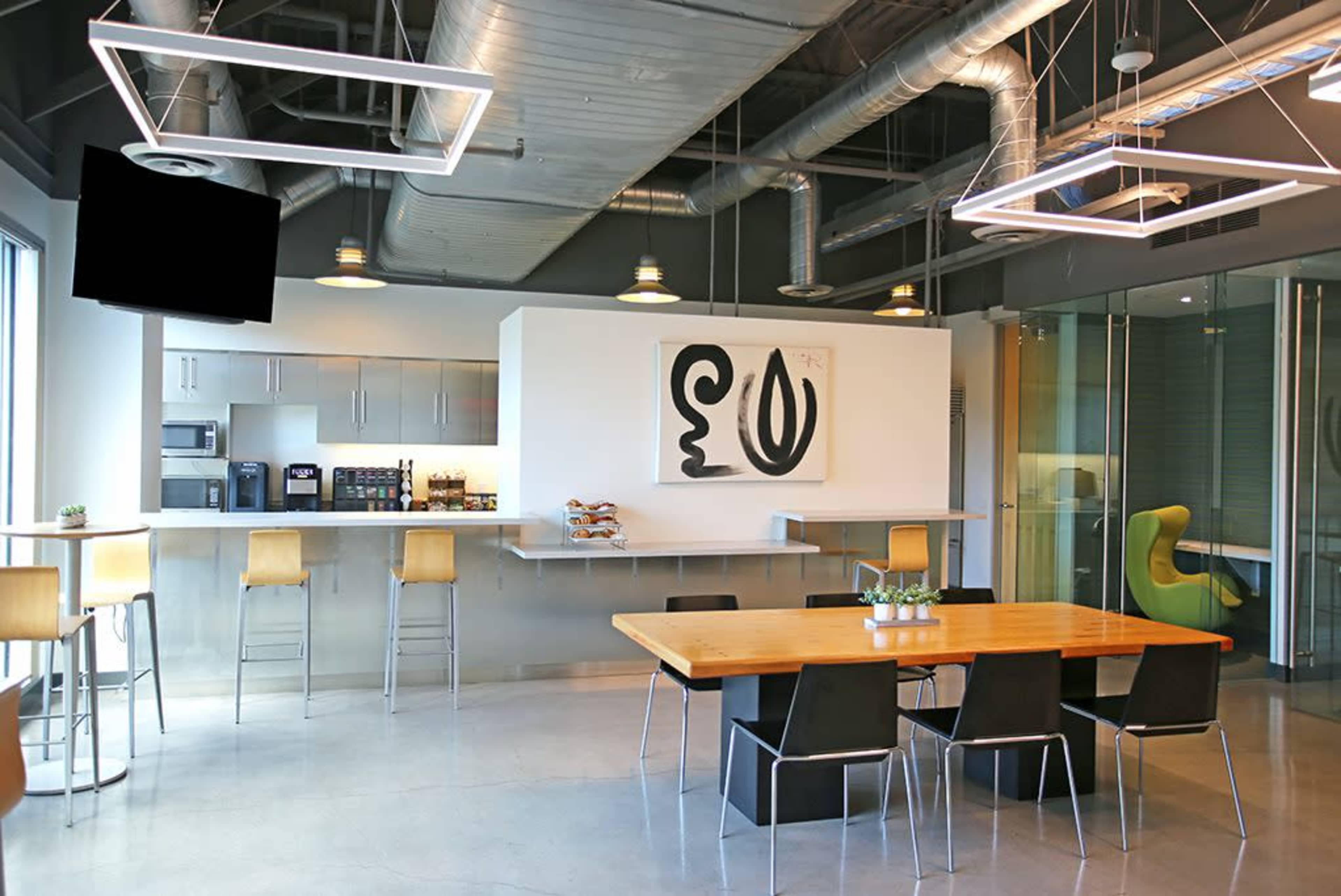 The image shows a modern office kitchen area with a long wooden table, black chairs, a bar with stools, and a coffee station against a backdrop of large windows and industrial-style decor.