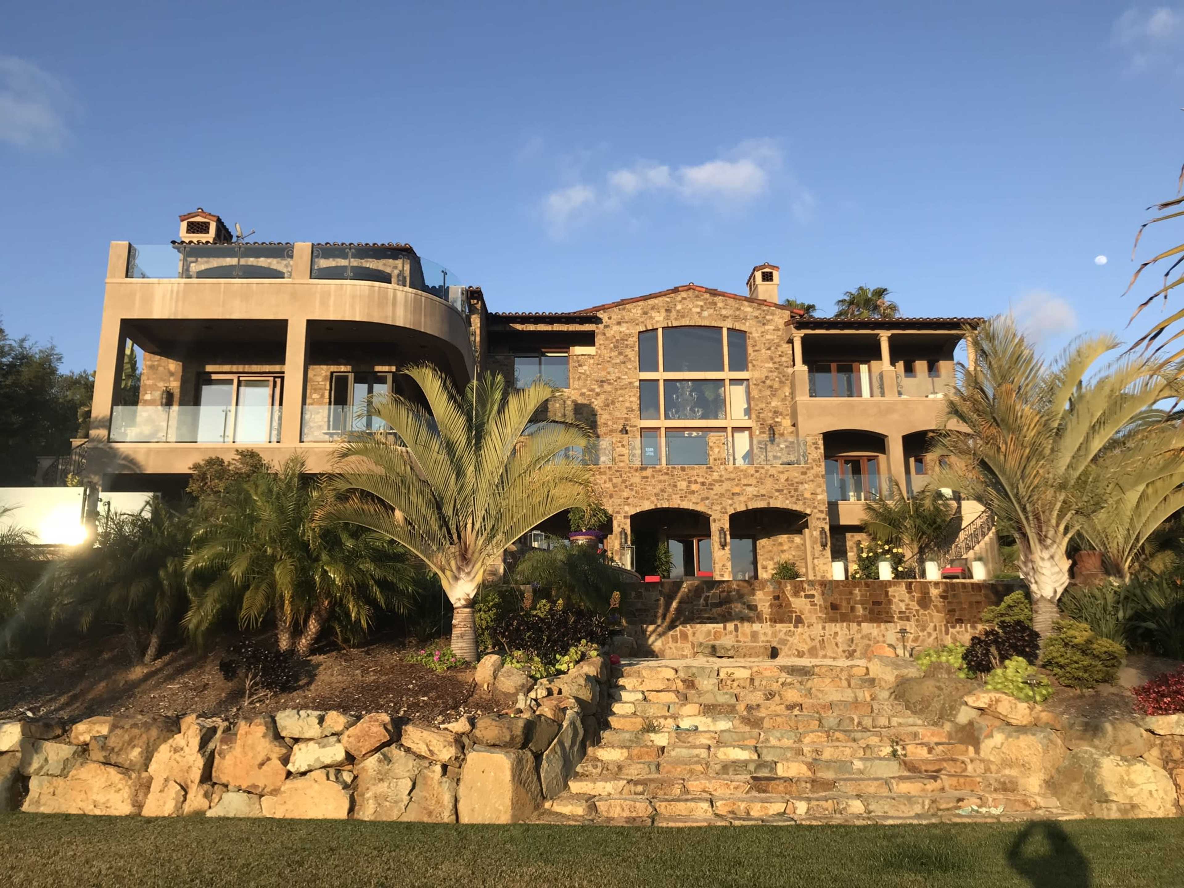 A large stone and concrete house with multiple levels, surrounded by palm trees and a stone staircase leading to the entrance.