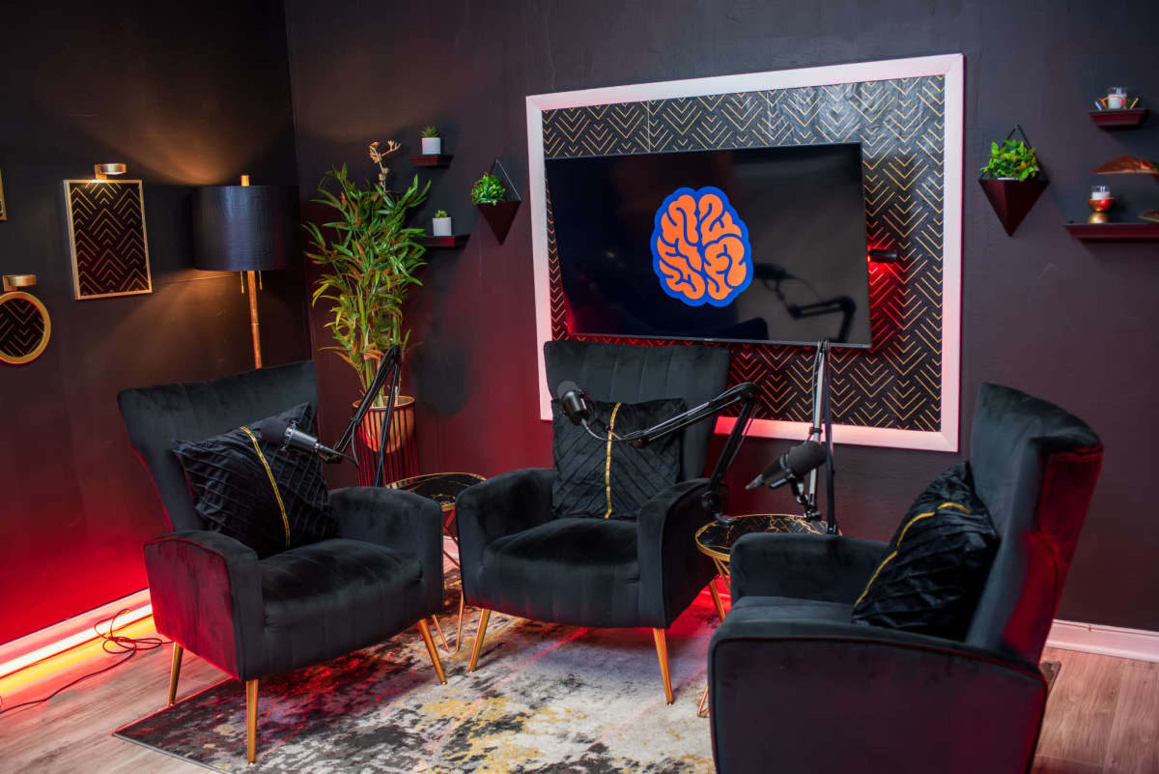 The image shows a modern discussion space with three black velvet chairs arranged around a wall-mounted TV displaying an illustrated brain logo, surrounded by decorative wall elements.