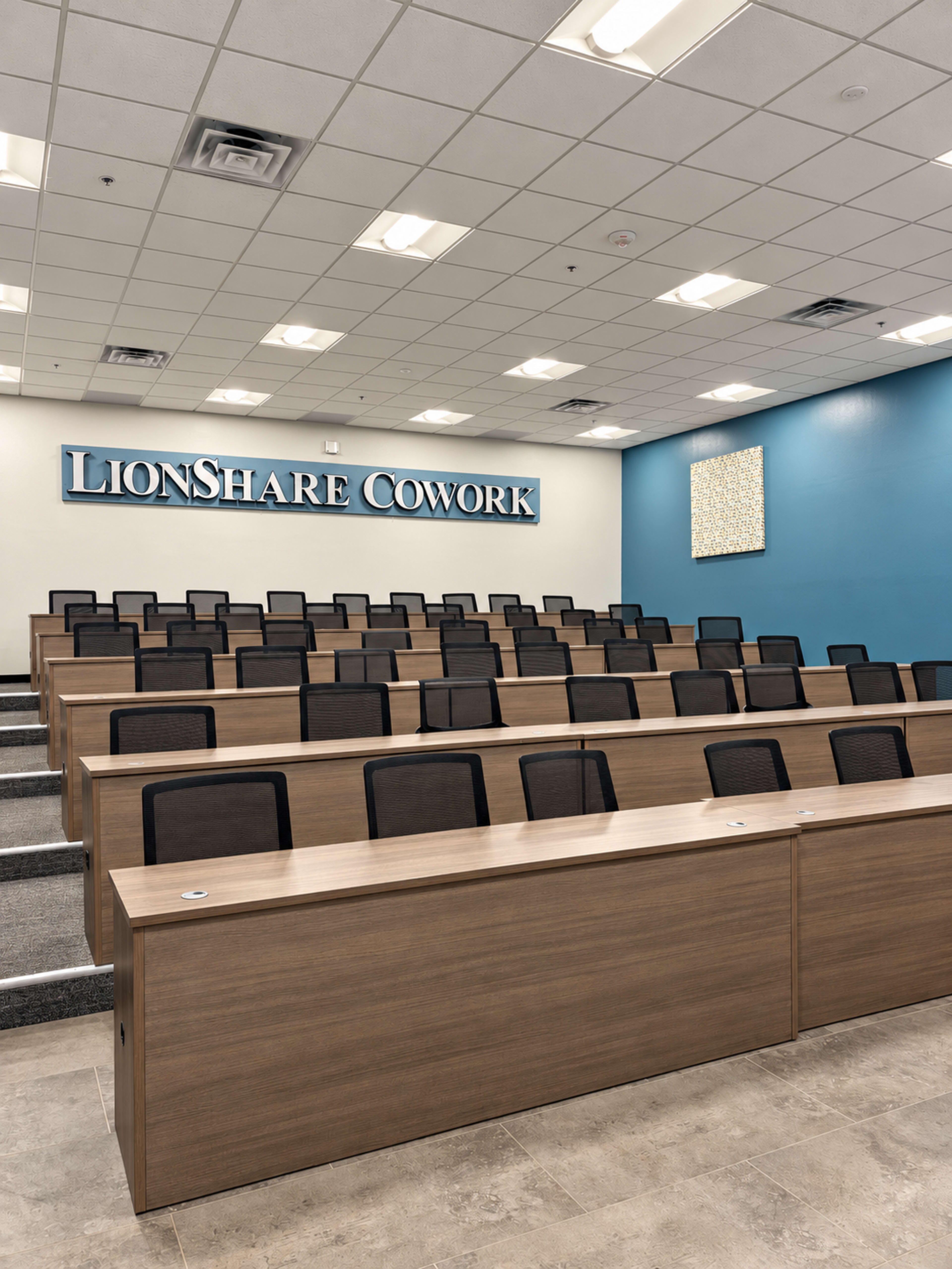 The image shows a classroom-style meeting space with tiered seating, wooden desks, and a wall featuring the text "LionShare Cowork."