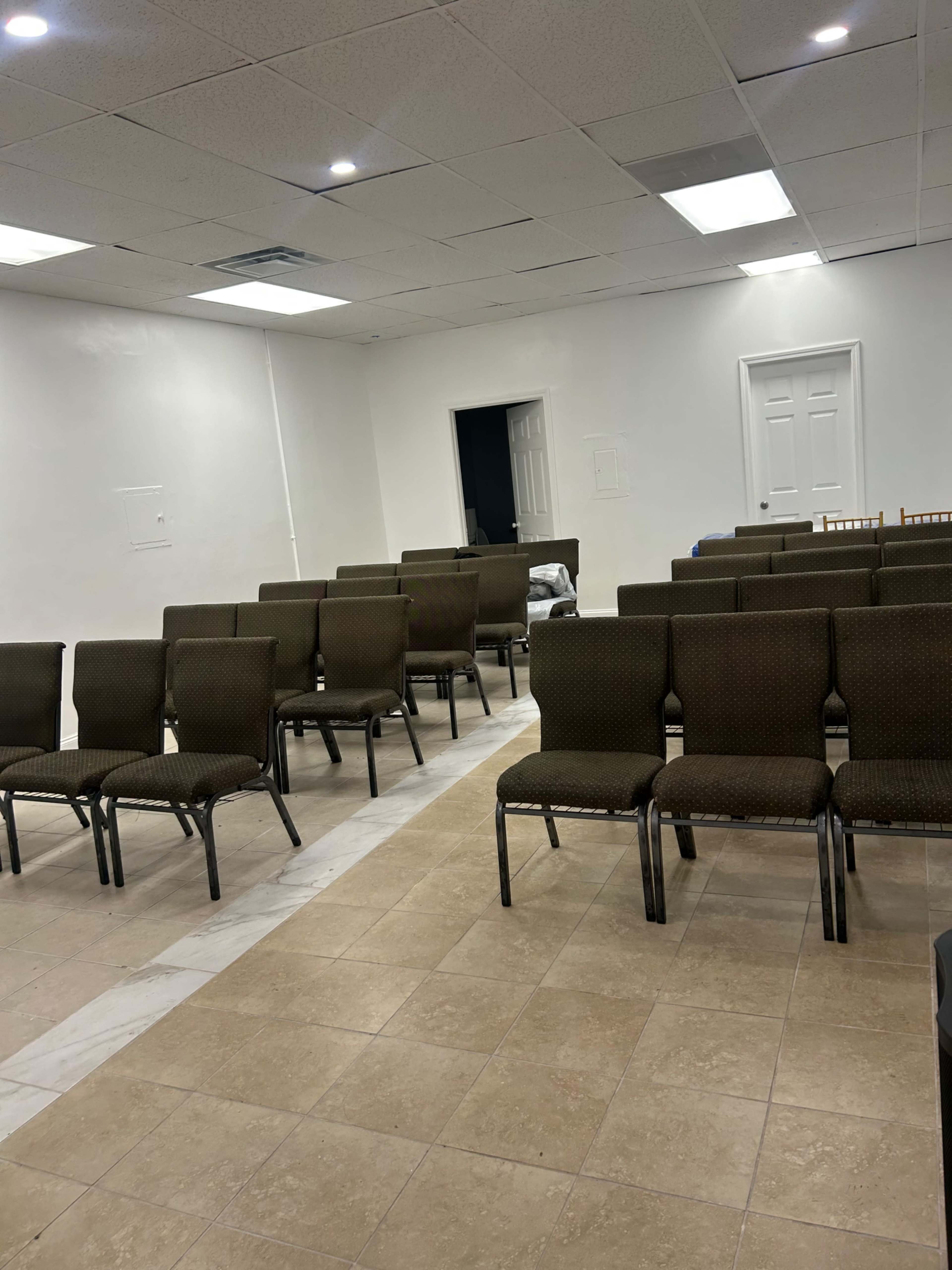 The image shows a room with several rows of brown chairs arranged on a tiled floor, with a doorway and unoccupied space in the background.