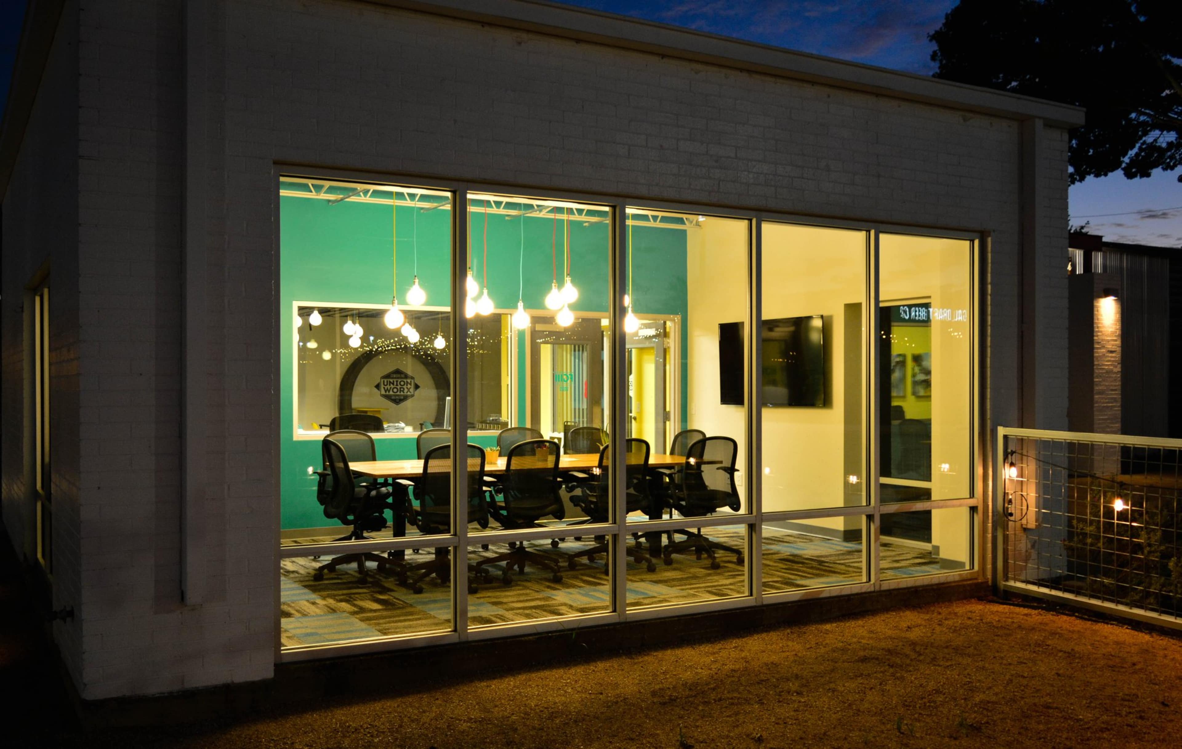 A modern conference room with glass walls features several black office chairs around a long table, illuminated by hanging lights.