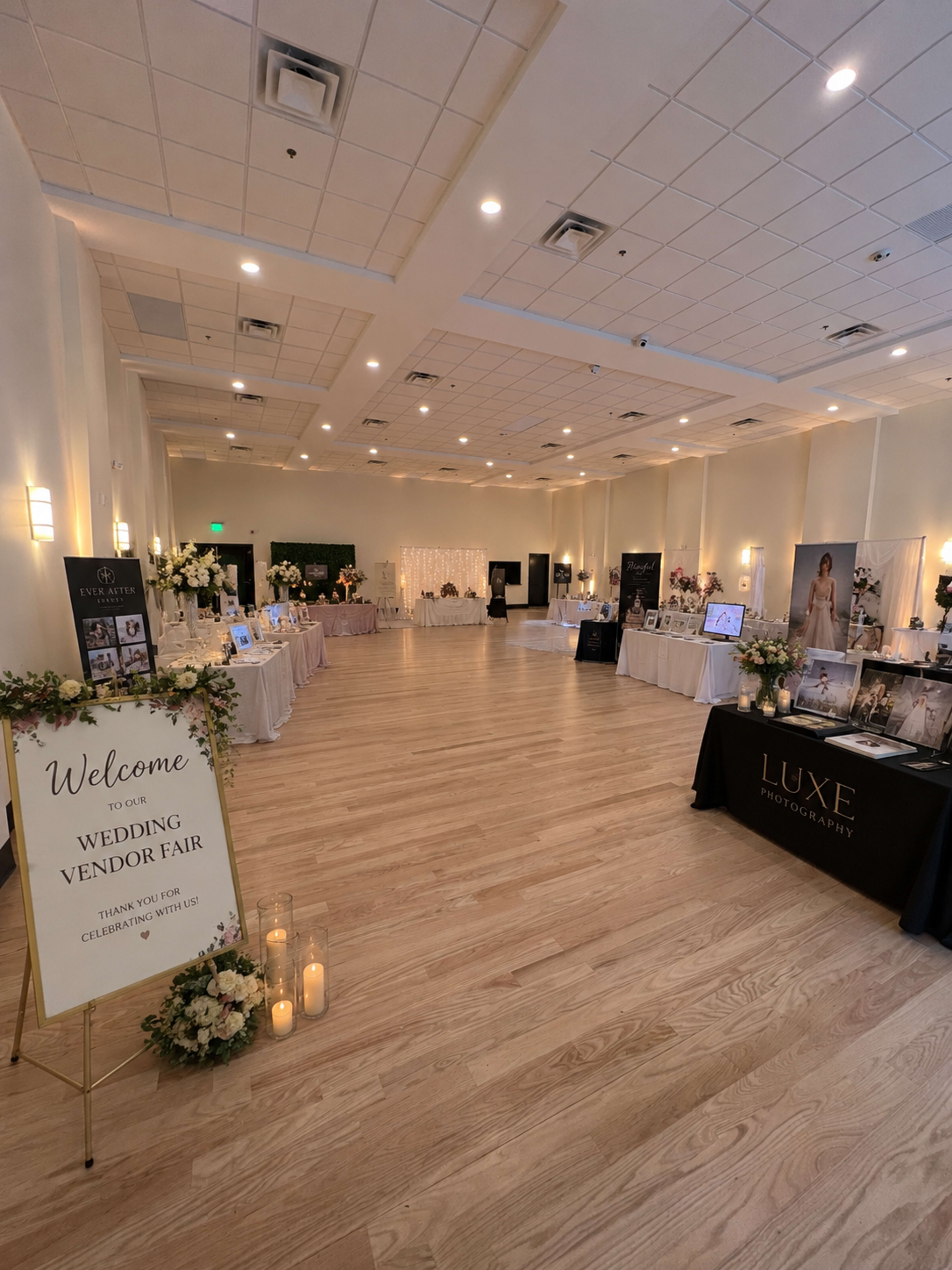 The image shows an indoor space set up for a wedding vendor fair, featuring several vendor tables displayed along the sides and a welcome sign at the entrance.