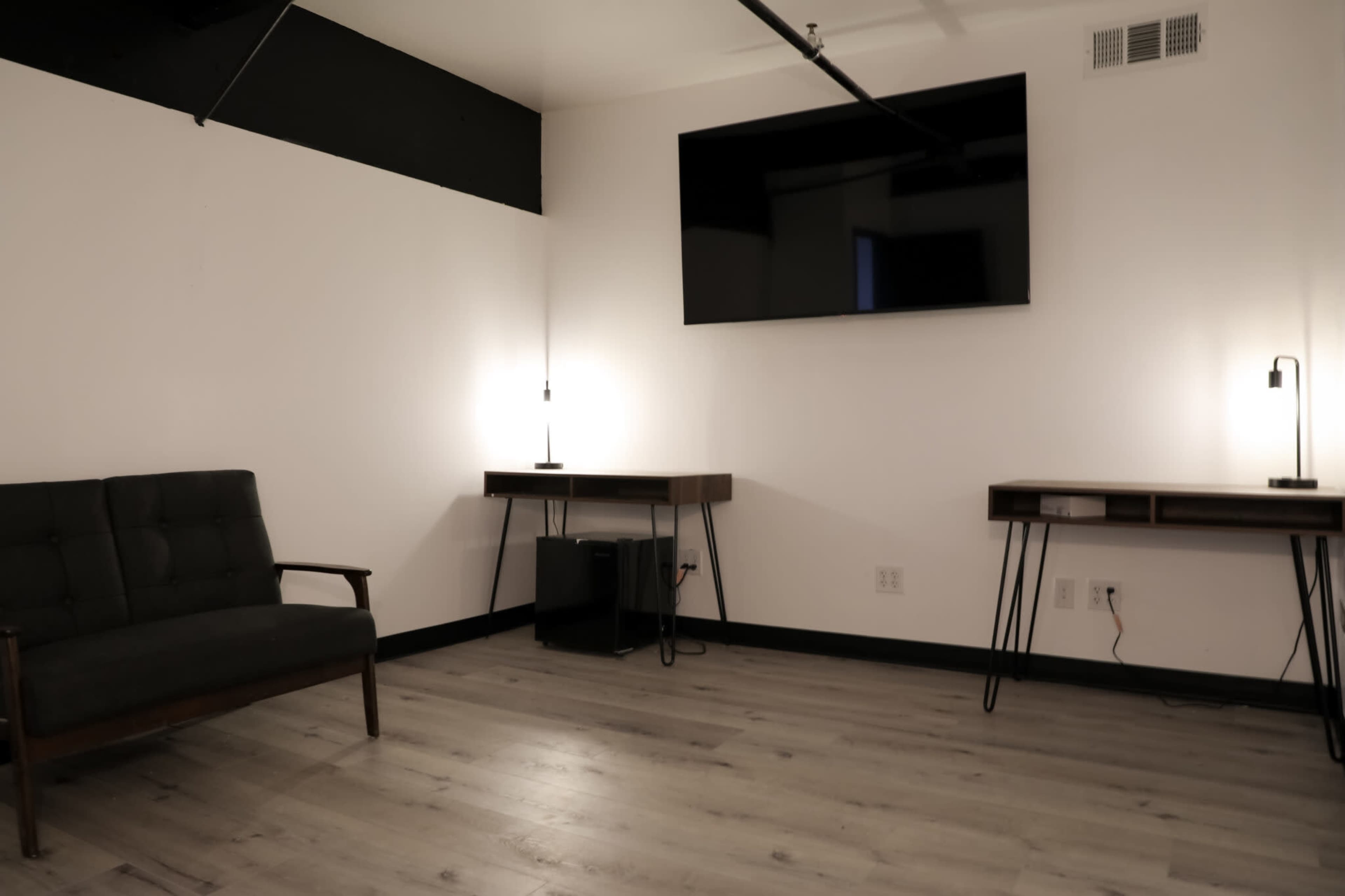 A minimalistic room with a dark sofa, two light fixtures on either side, and a wall-mounted television above a pair of wooden desks.