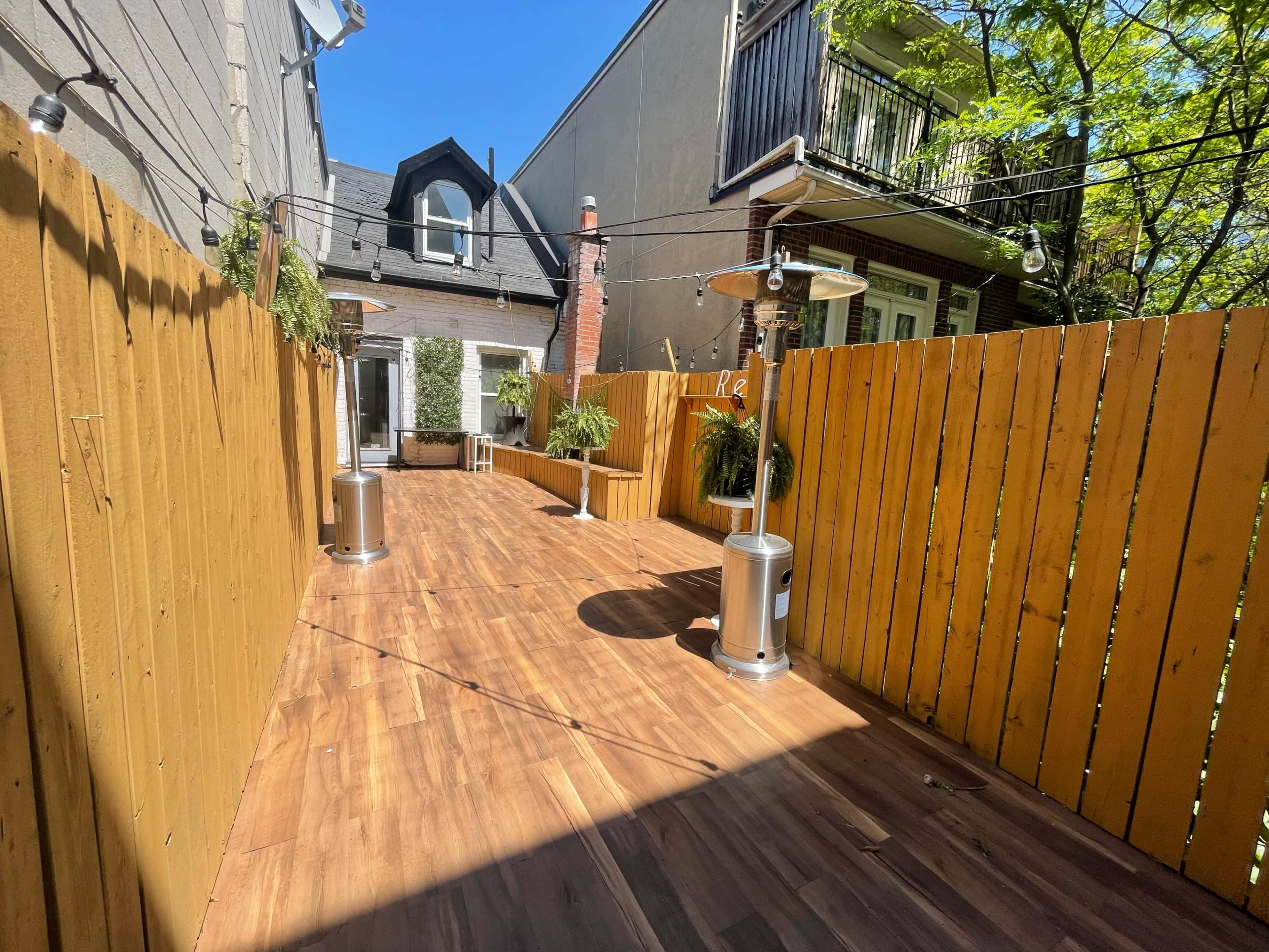 The image shows a wooden deck area enclosed by a tall wooden fence, featuring patio heaters and greenery along the sides.