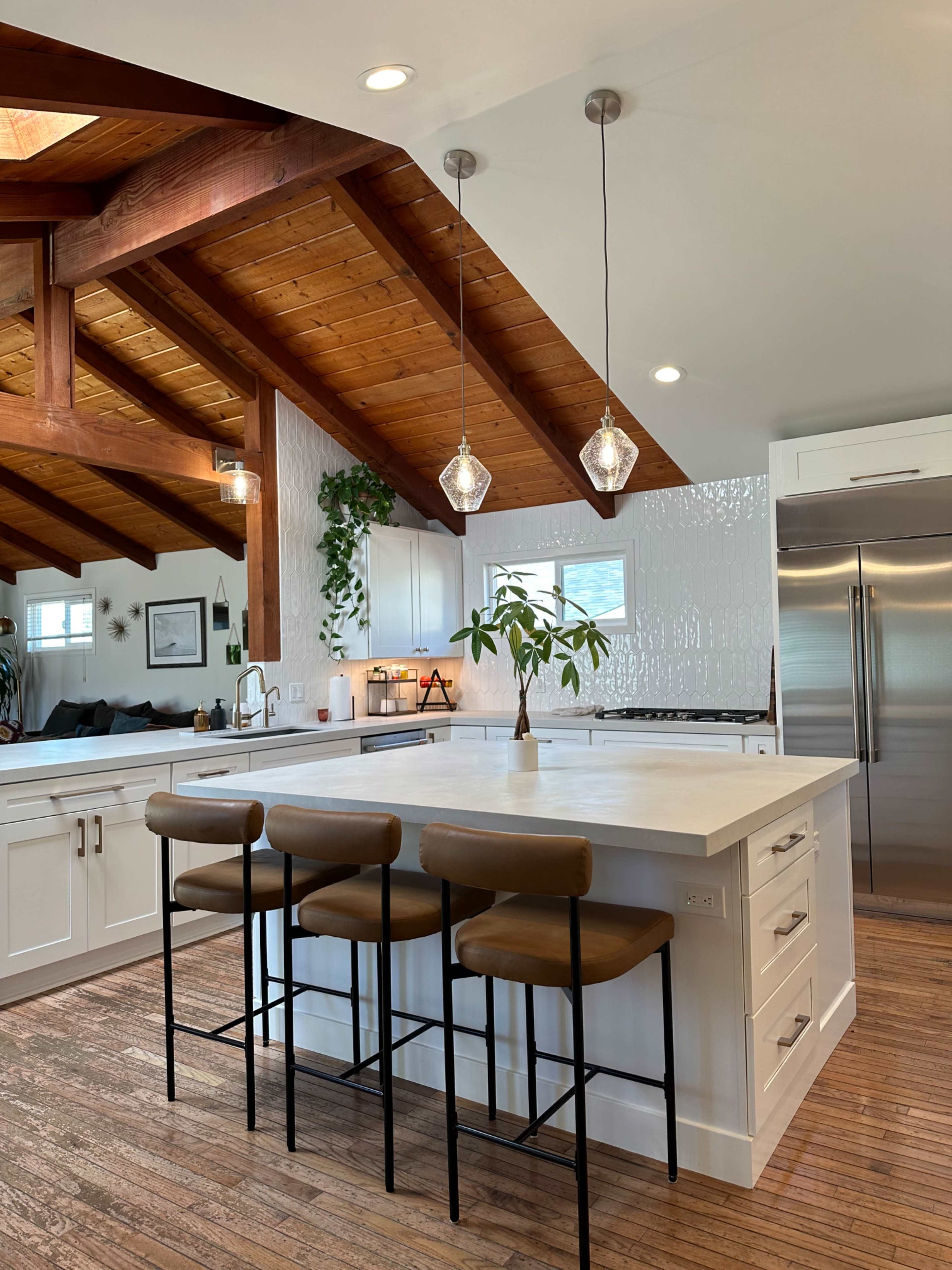 A modern kitchen with a large island, featuring white cabinetry, wooden beams, and three bar stools.