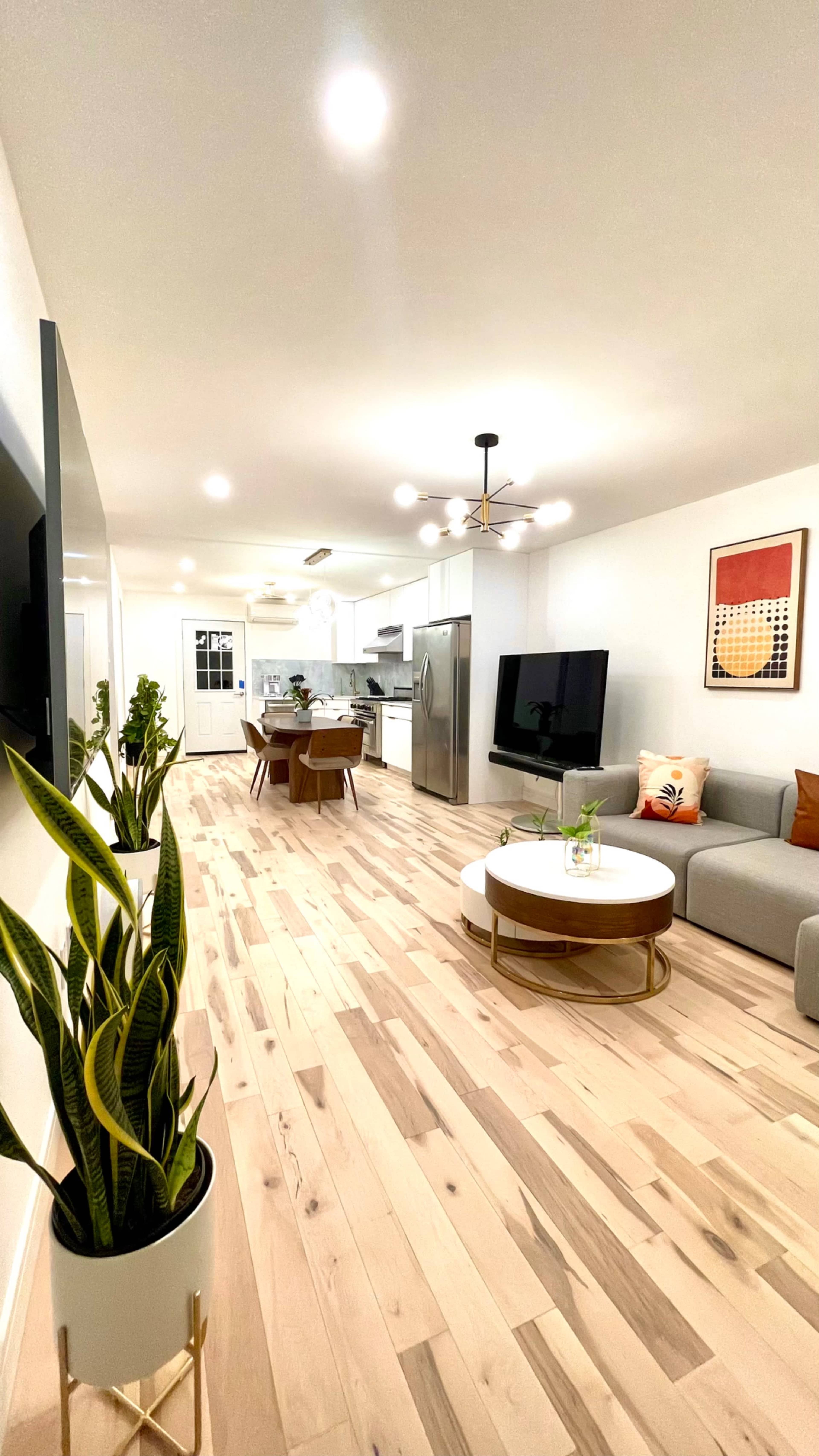 The image shows a modern living room with light wooden flooring, a gray couch, and a round coffee table, leading to an open kitchen area with dining space.