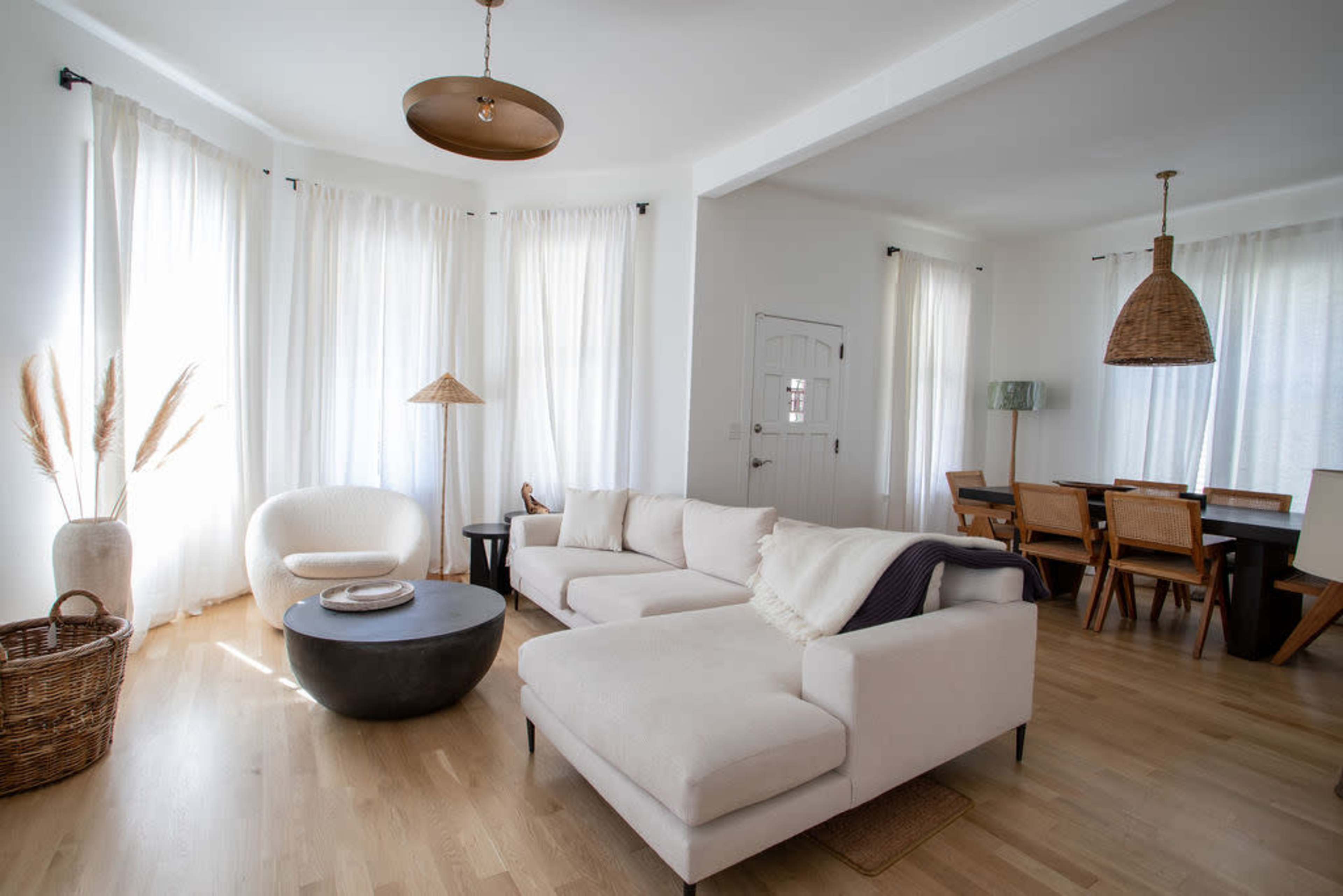 A bright living room with a light-colored sectional sofa, a round coffee table, large windows draped in white curtains, and a dining area with wooden chairs and a table.