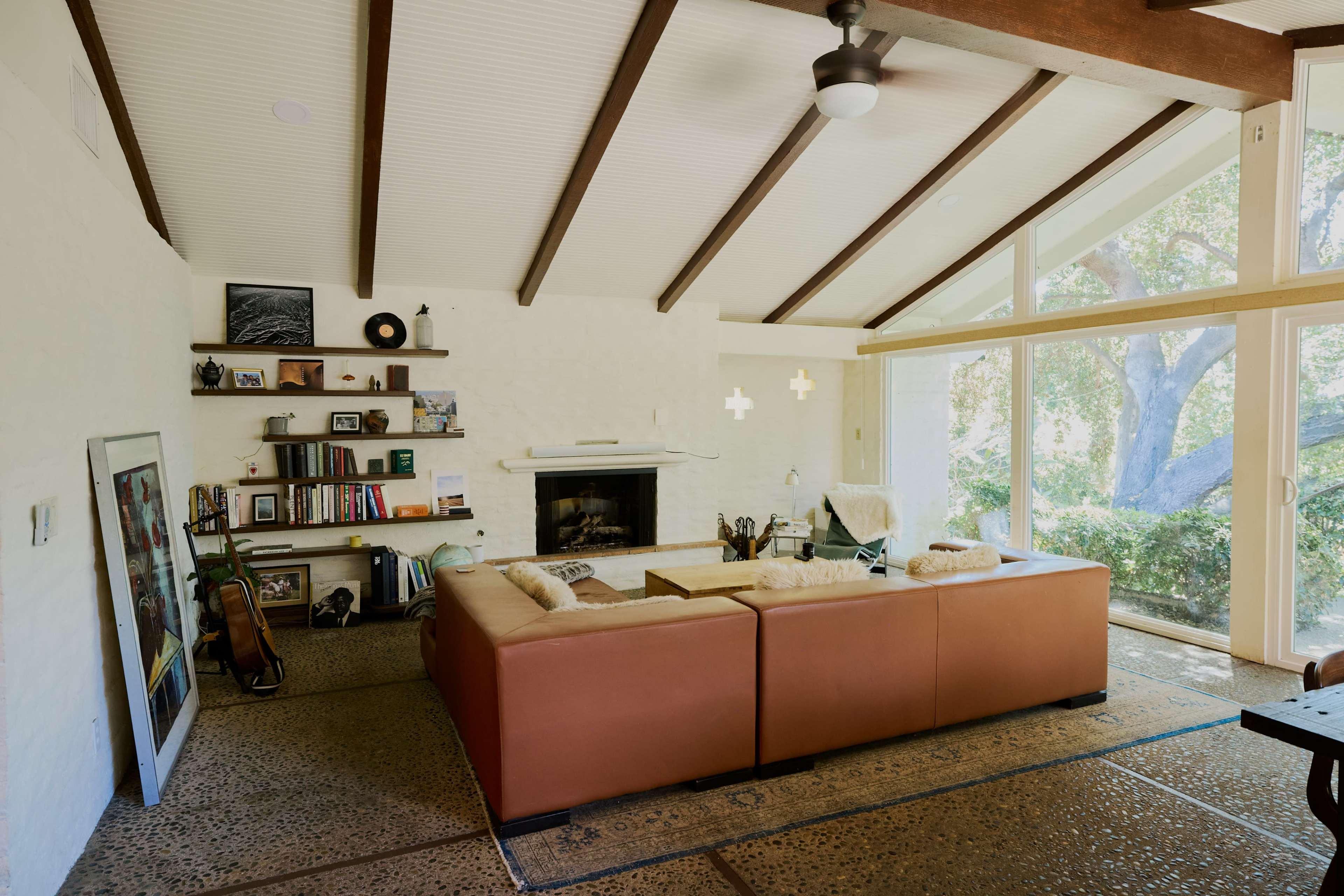 A modern living room features a brown sectional sofa, a fireplace, built-in shelves with books, and large windows overlooking a tree-filled view.
