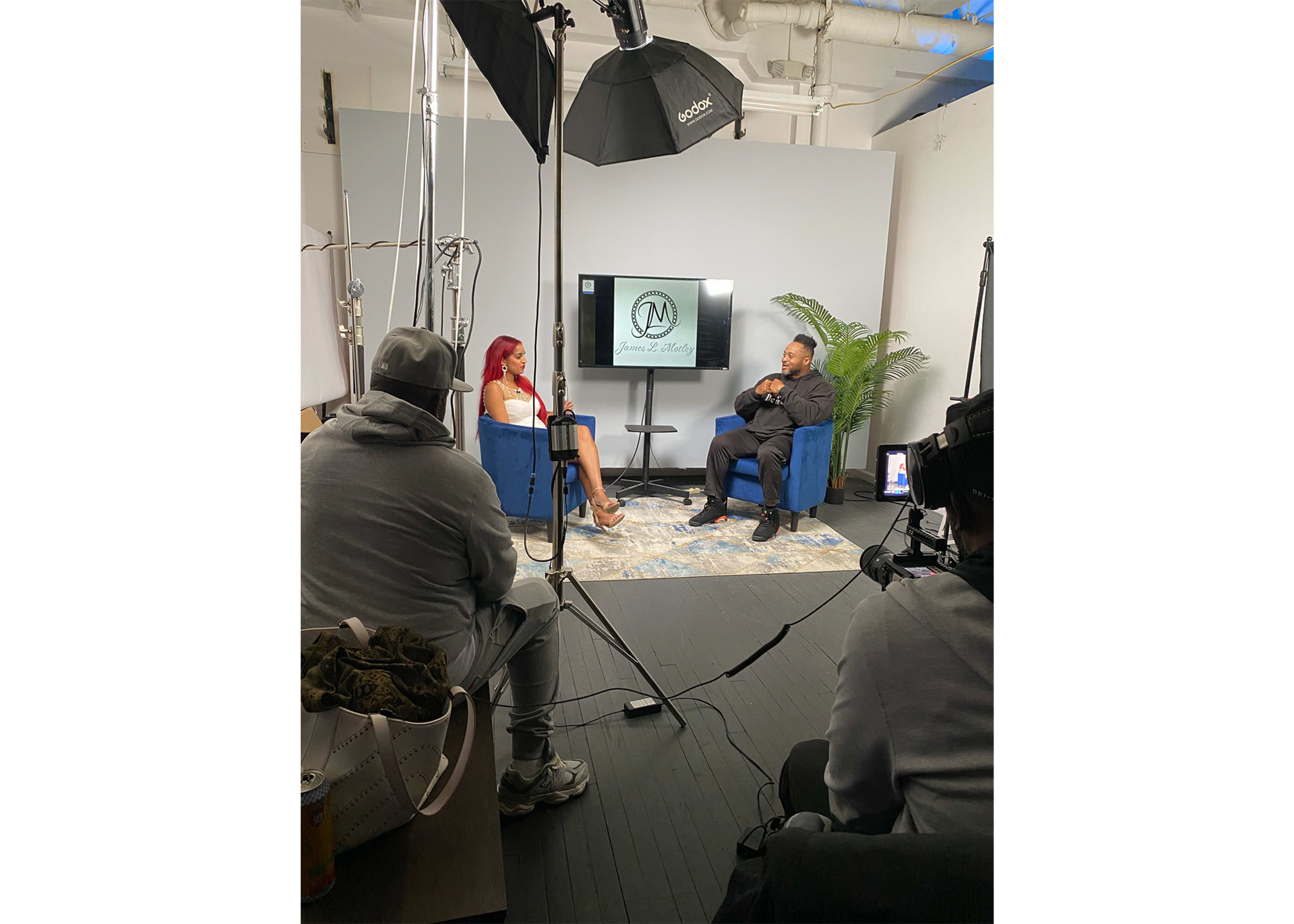 A woman and a man are seated in blue chairs during an interview in a studio setting with a camera crew and lighting equipment.