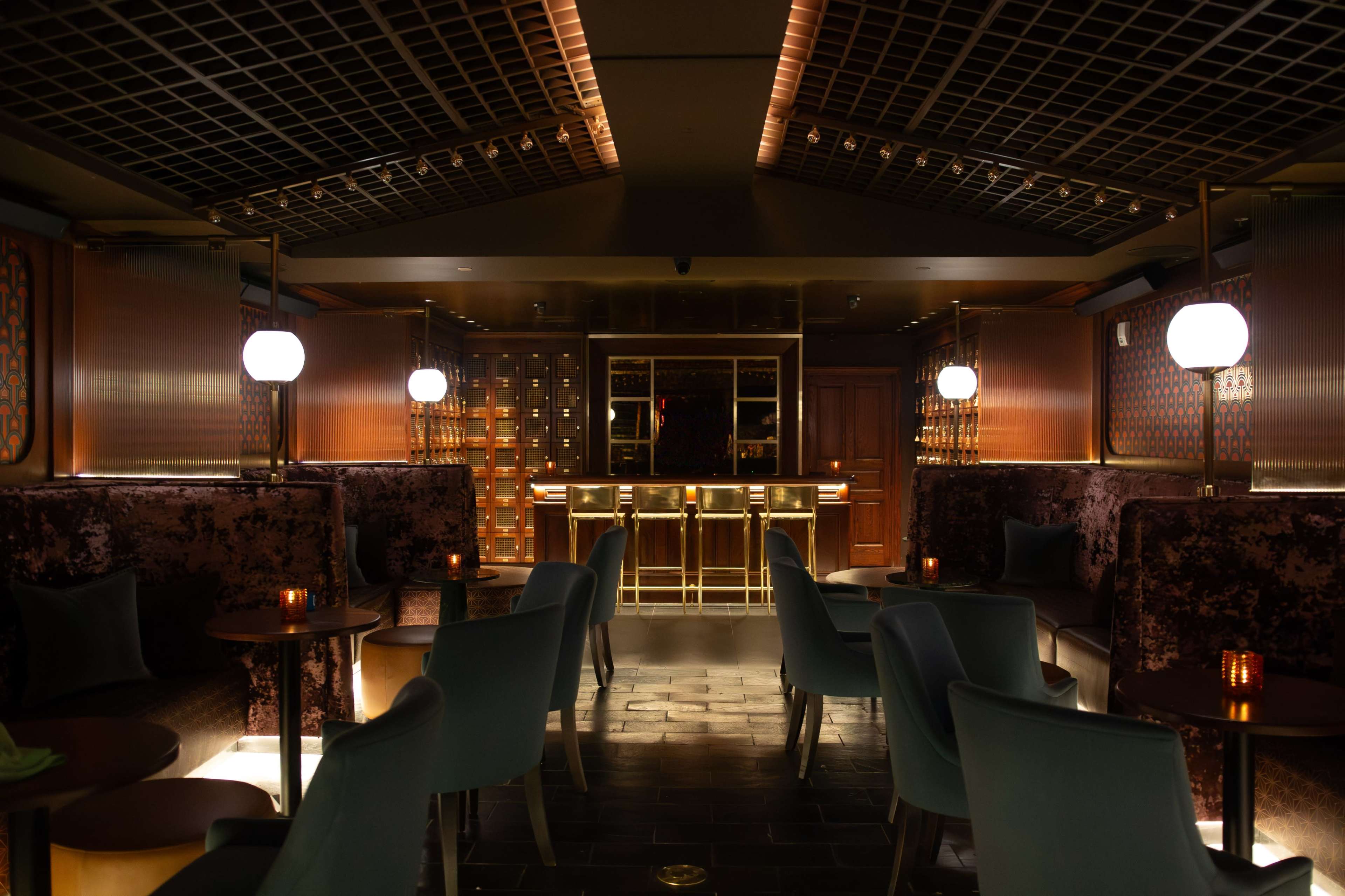 The image shows a dimly lit, elegant bar with plush seating, a glowing counter, and decorative lighting fixtures.