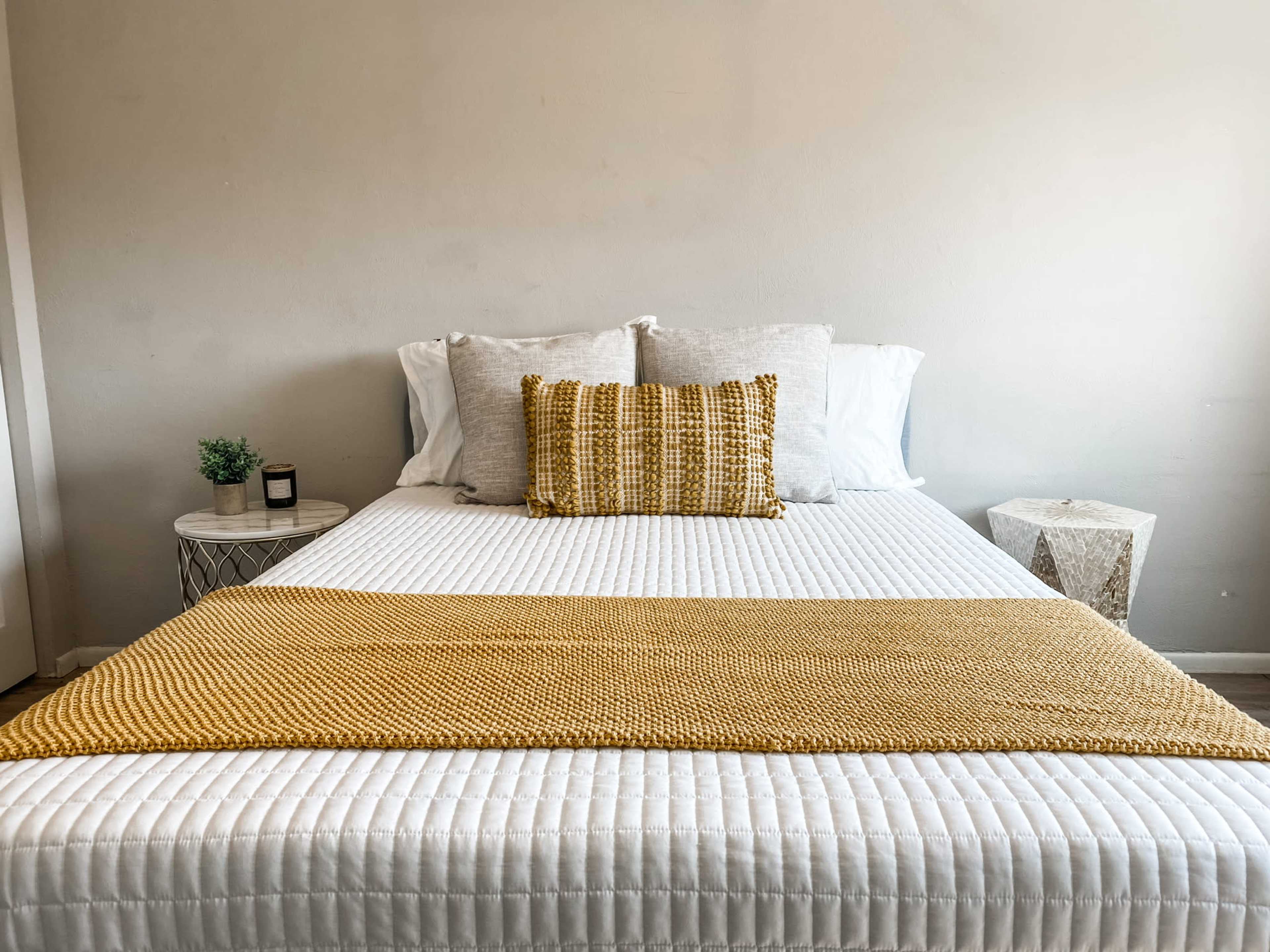 A neatly made bed with a white quilted cover, decorative pillows, and a yellow throw blanket is positioned against a neutral wall, accompanied by a small plant and a geometric side table.