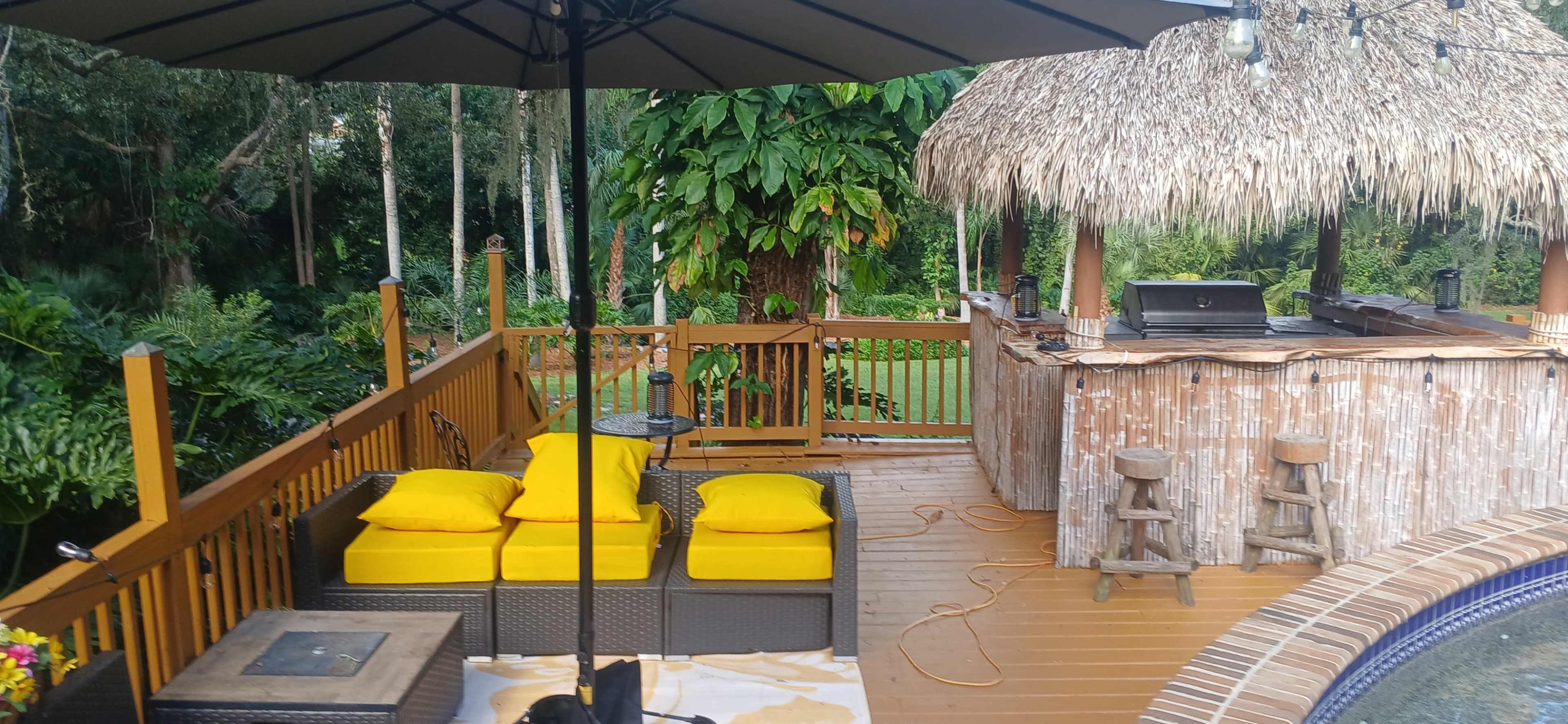 The image shows a shaded outdoor patio area with yellow cushions on dark furniture, adjacent to a thatched bar and surrounded by lush greenery.