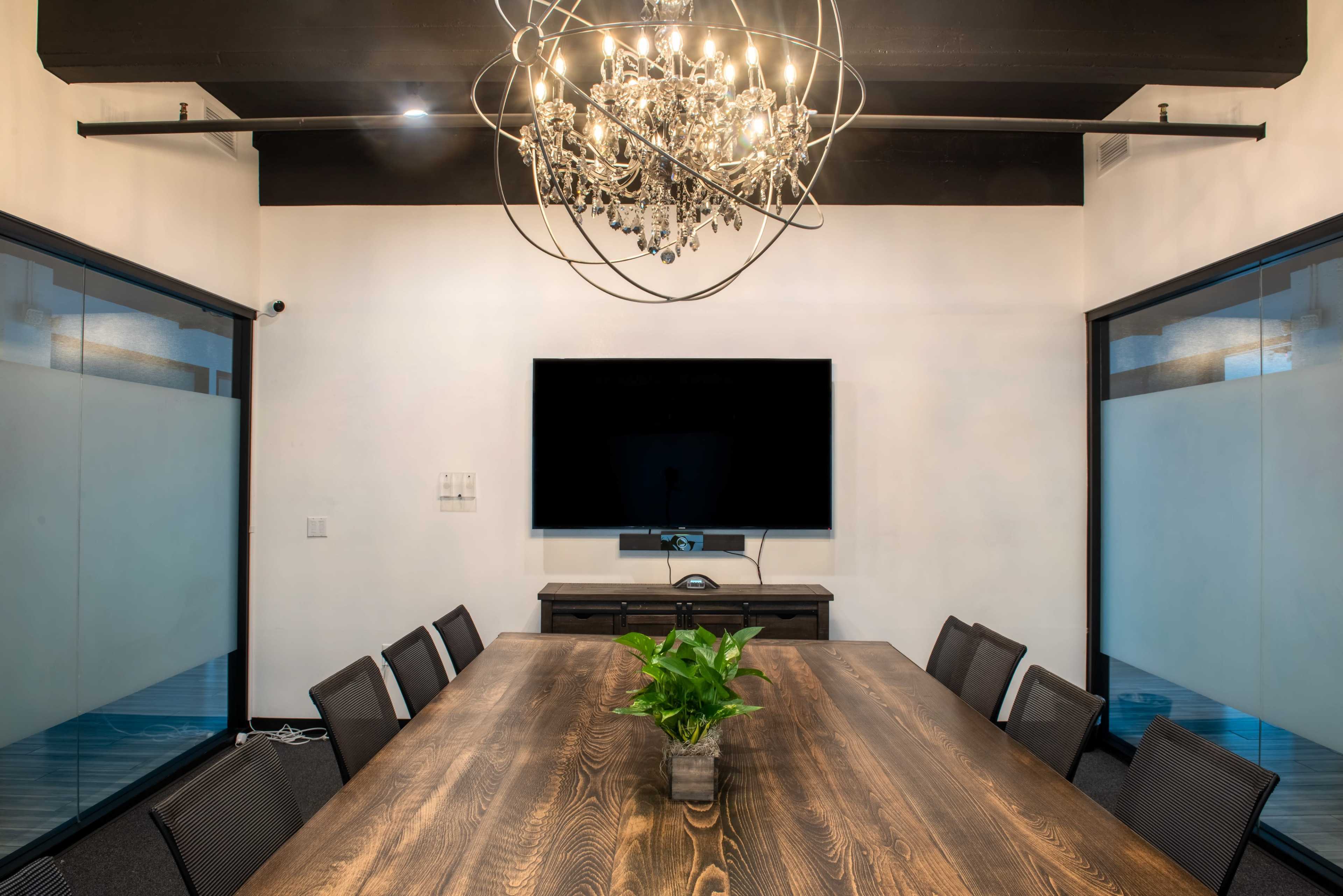 A long wooden conference table is set in a modern meeting room with a chandelier above and a large TV mounted on the wall.