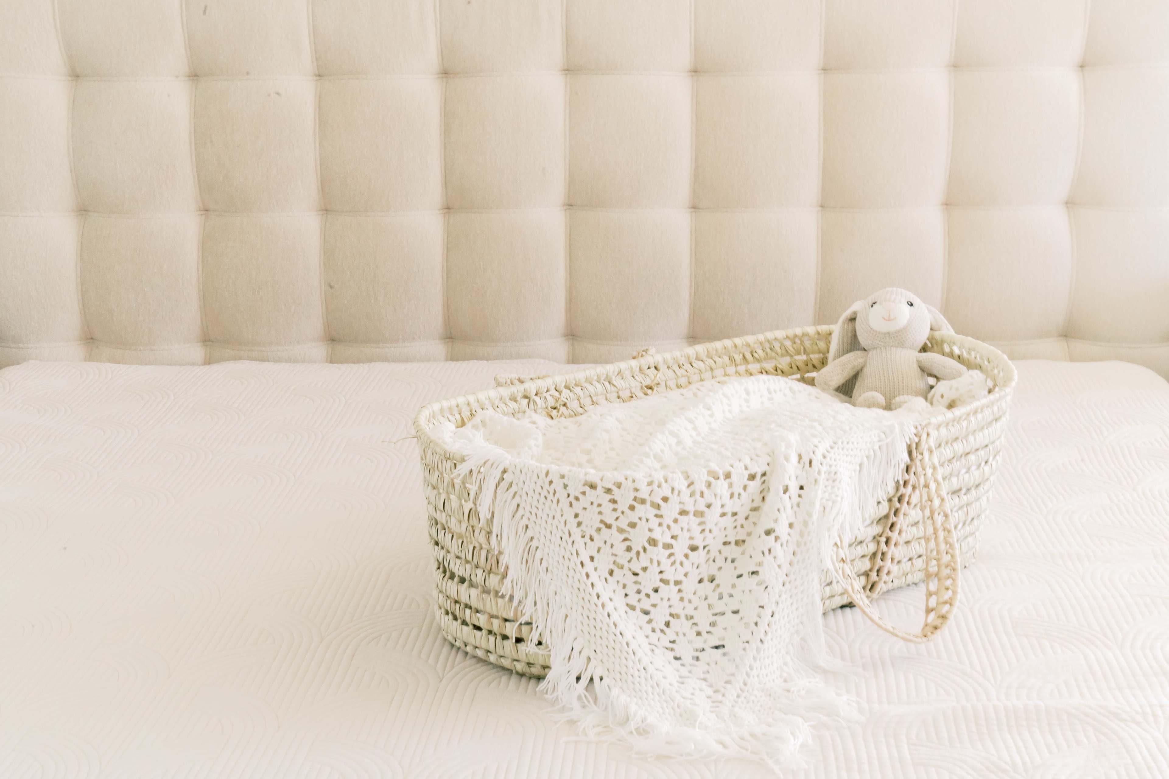A wicker basket lined with a blanket and a stuffed toy rabbit sits on a bed with a tufted headboard.