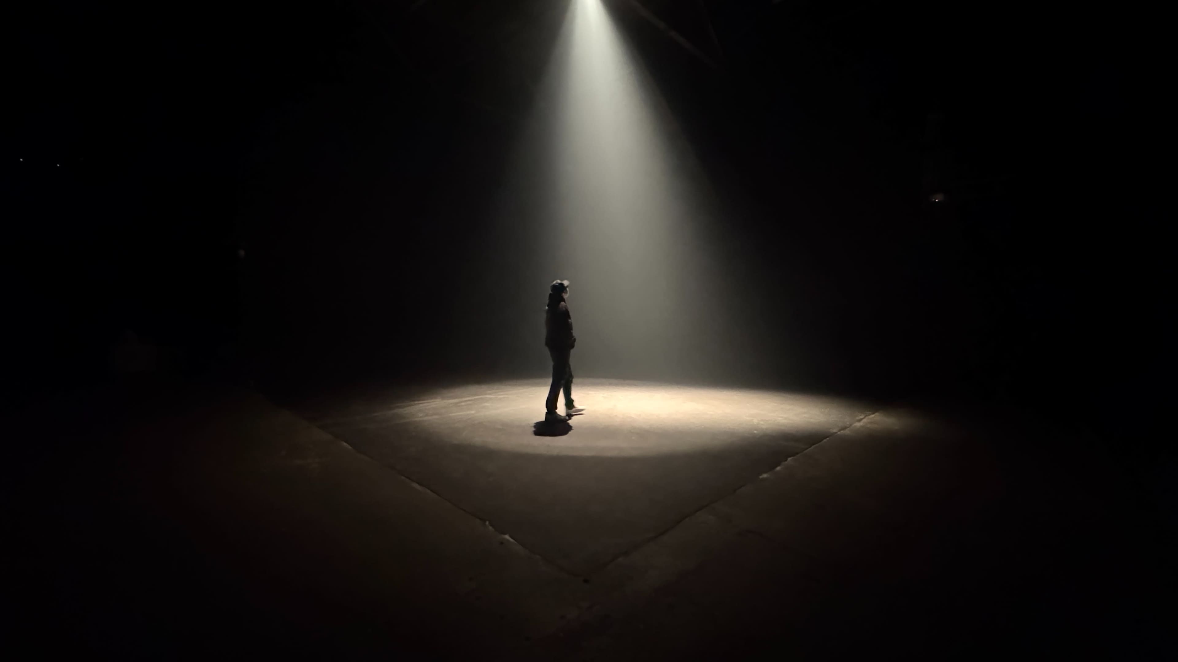 A solitary figure stands in a beam of light on a dark stage.