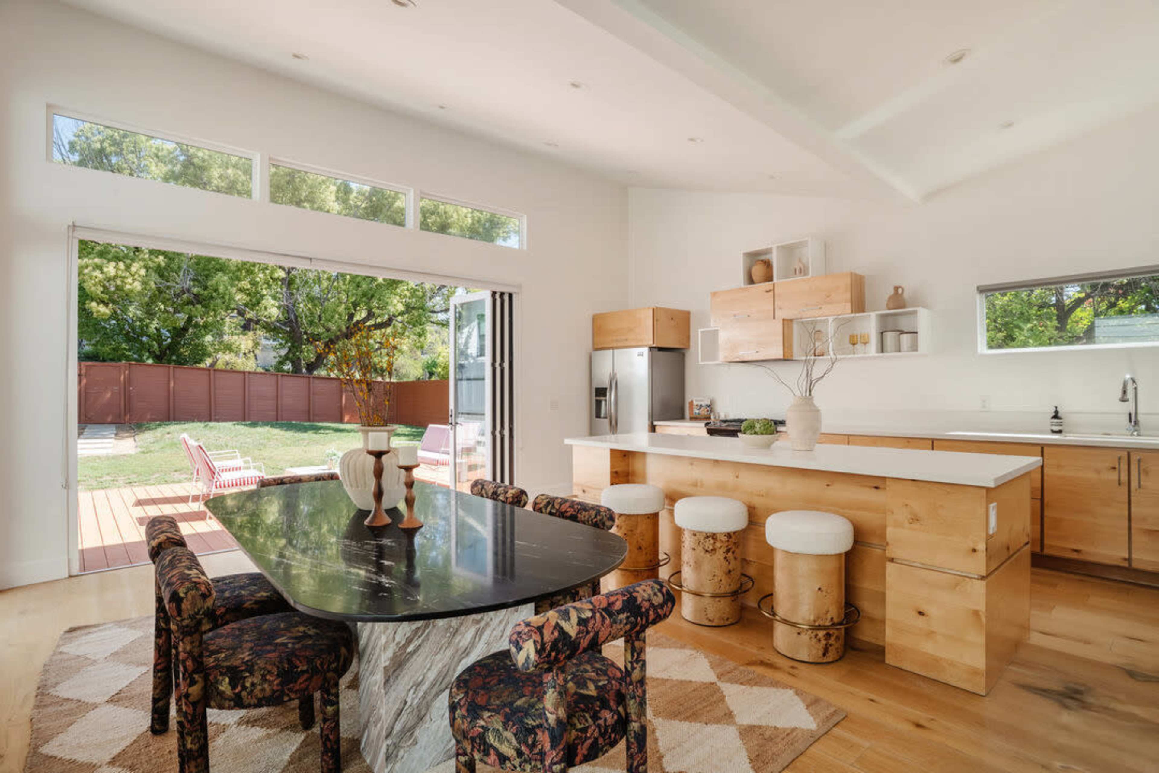 A modern kitchen features a black marble dining table, wooden cabinets, and large windows that provide a view of the outdoor space.