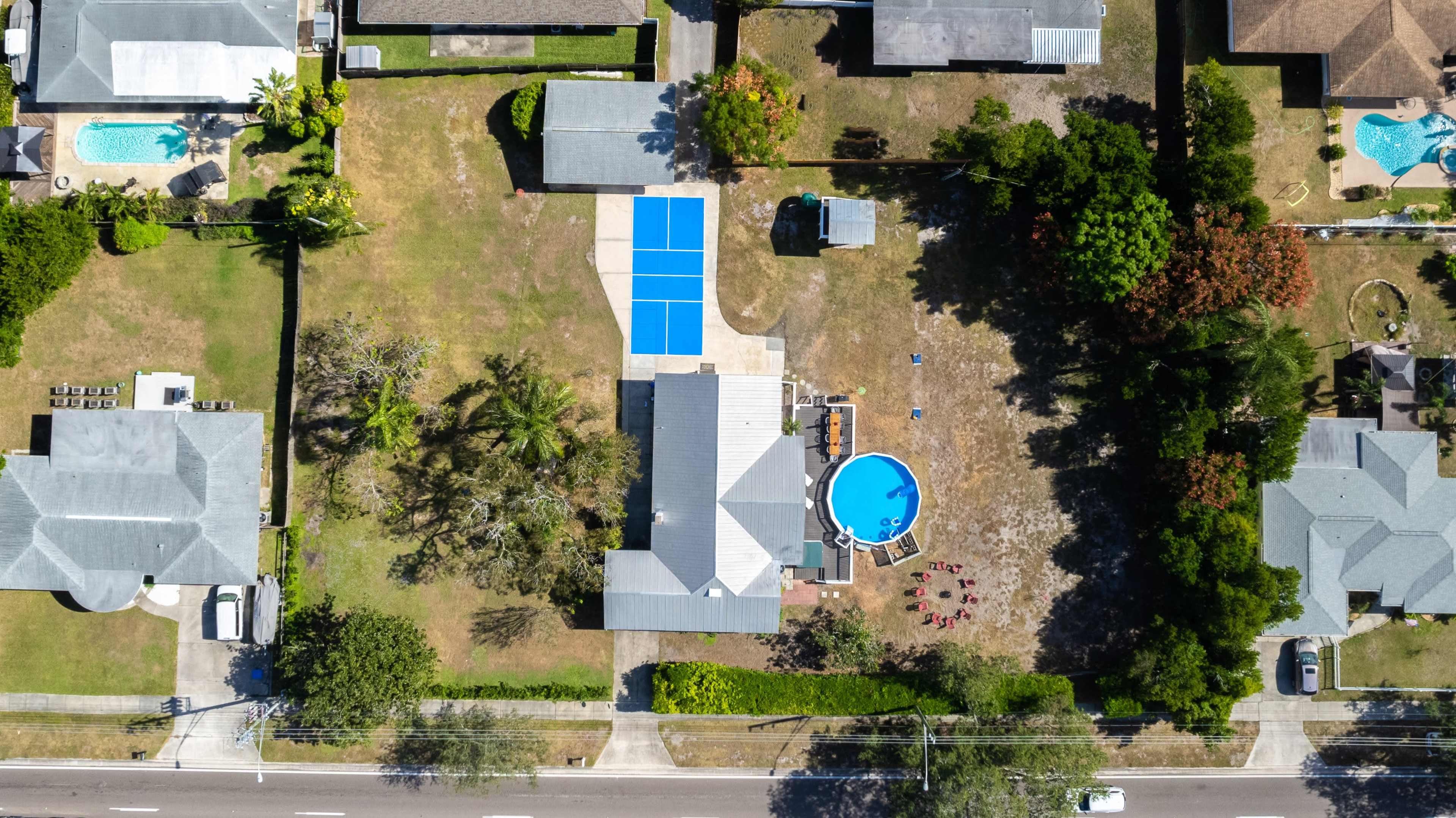 An aerial view of a residential area featuring several houses, a pool in one backyard, and an organized layout of blue paddle courts nearby.