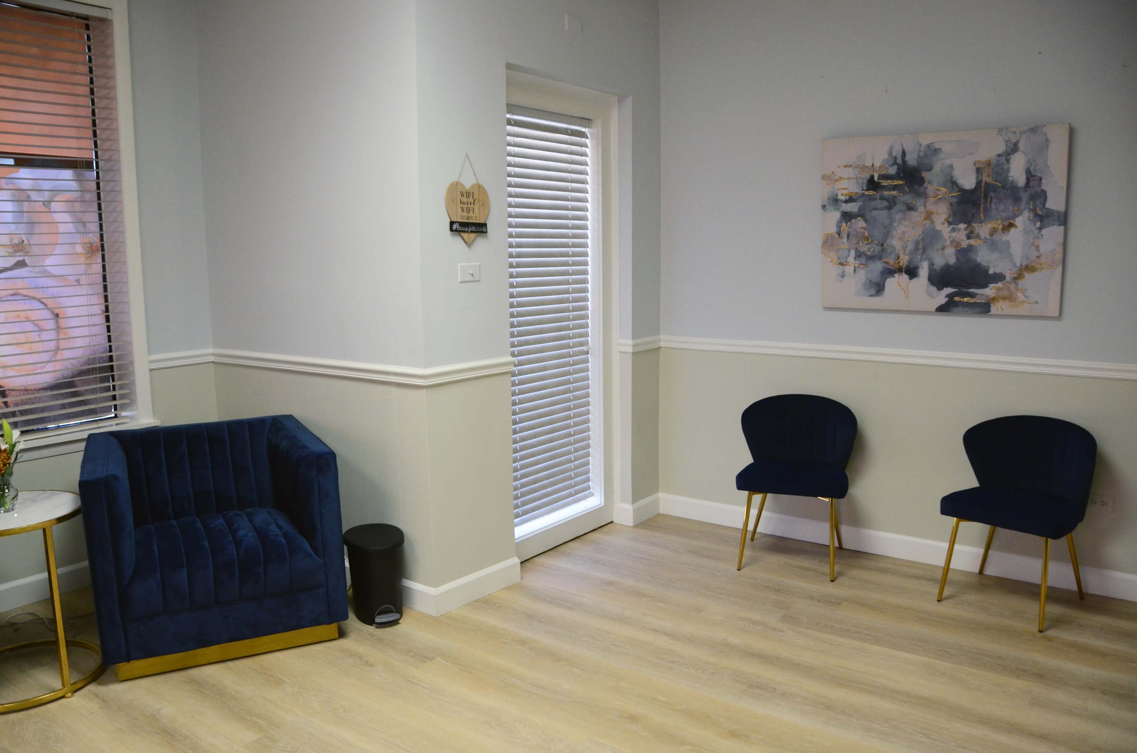 A minimally furnished room with a blue velvet armchair, two complementary chairs, and a door leading outside, framed by a light-colored wall and wooden flooring.