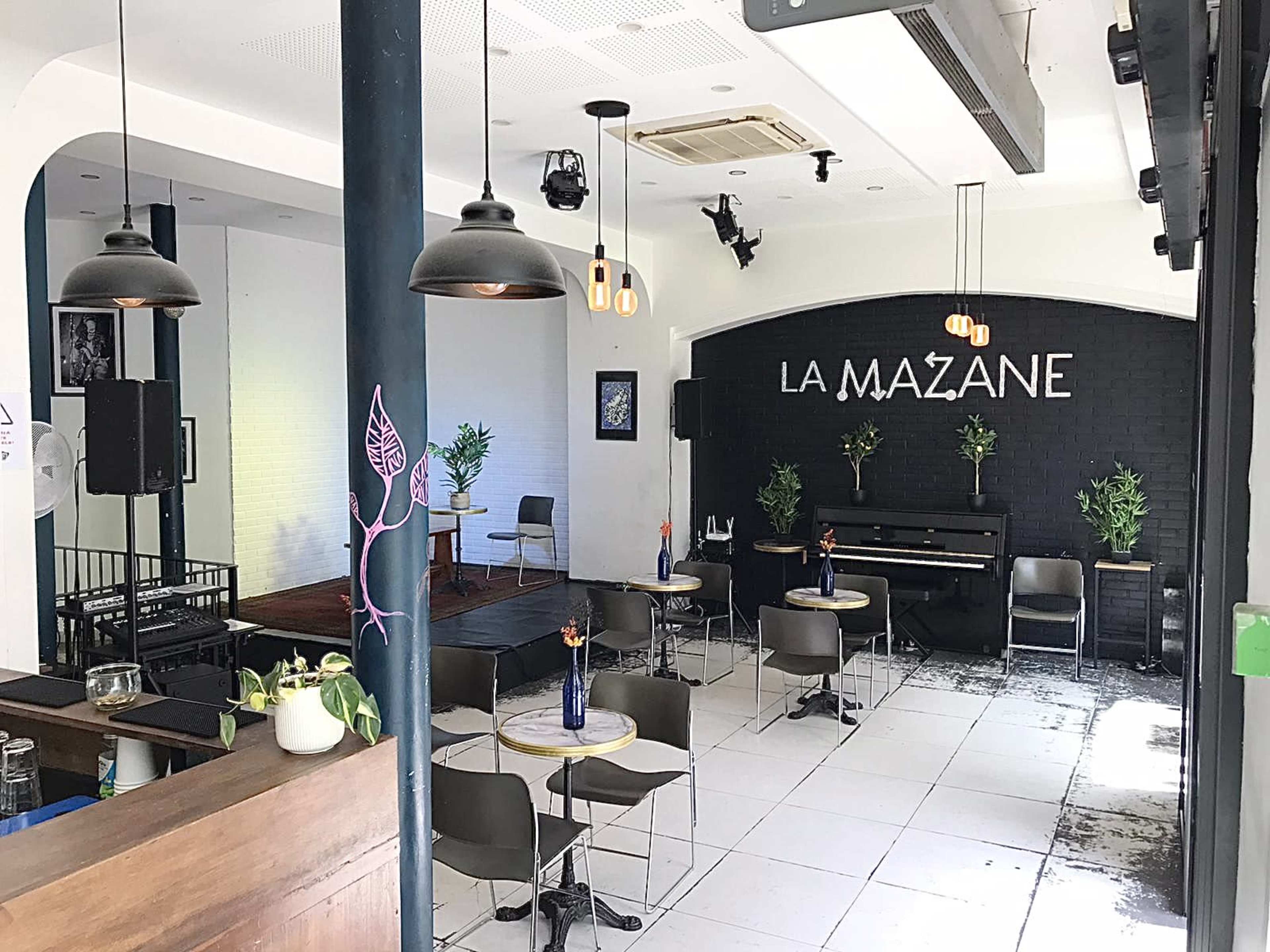 The image shows a modern café interior with seating arrangements, a piano, and a decorative wall featuring the name "LA MAZANE."