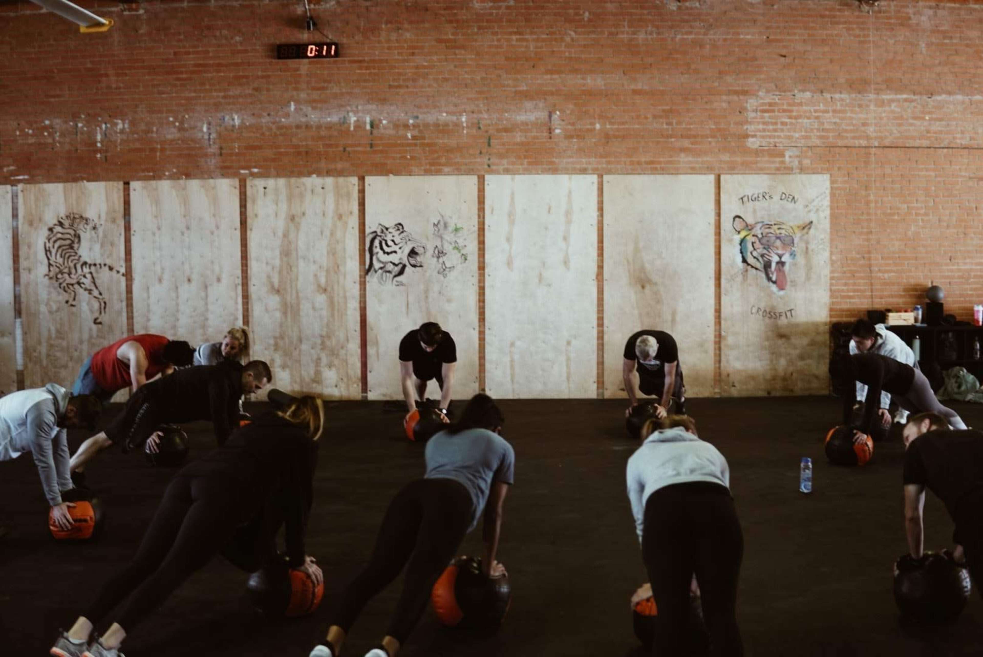 A group of people is performing a workout on a gym floor, using weighted balls while facing down in a plank position.