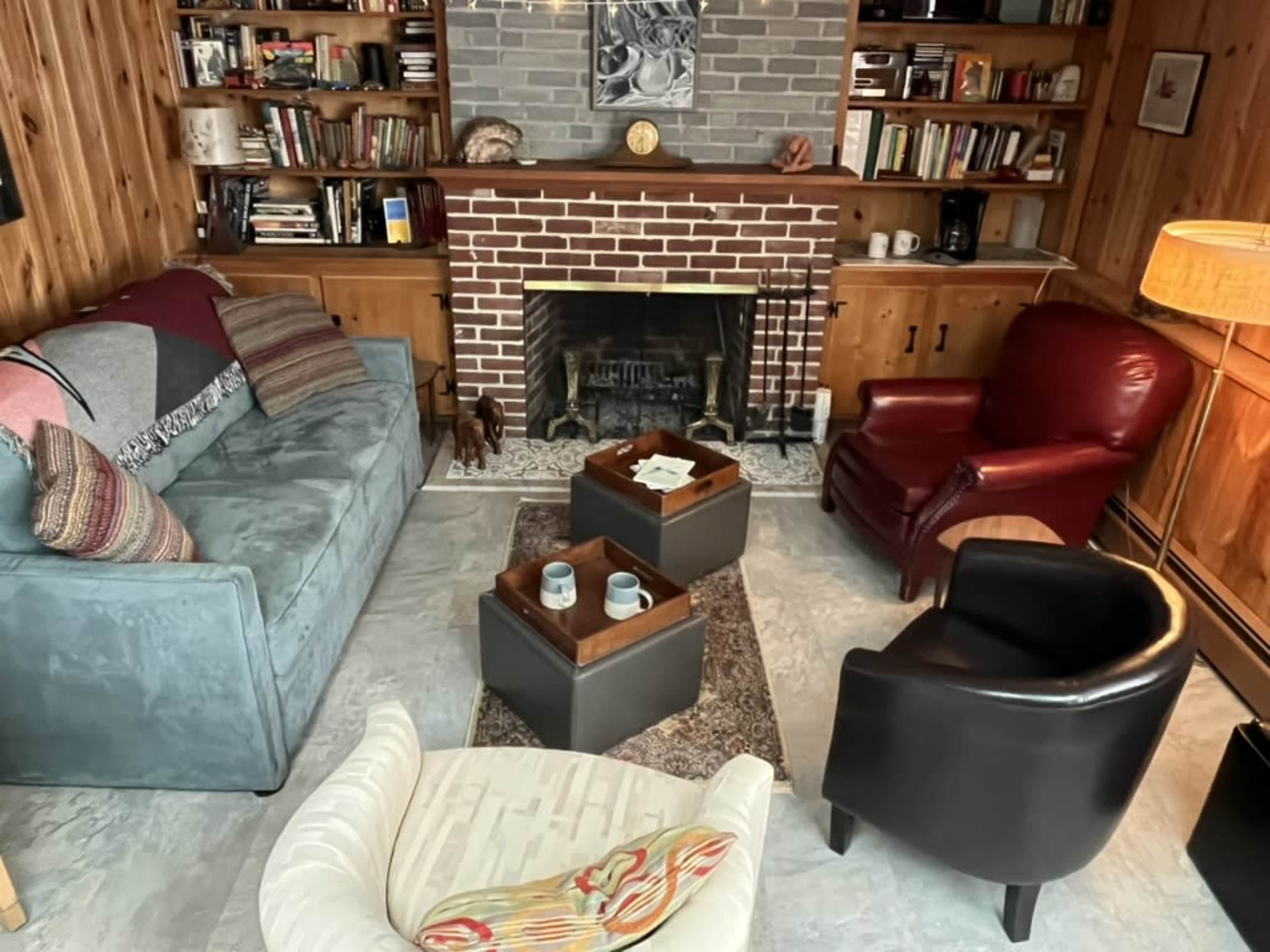 The image shows a cozy living room with a brick fireplace, surrounded by bookshelves, comfortable seating, and a coffee table in the center.