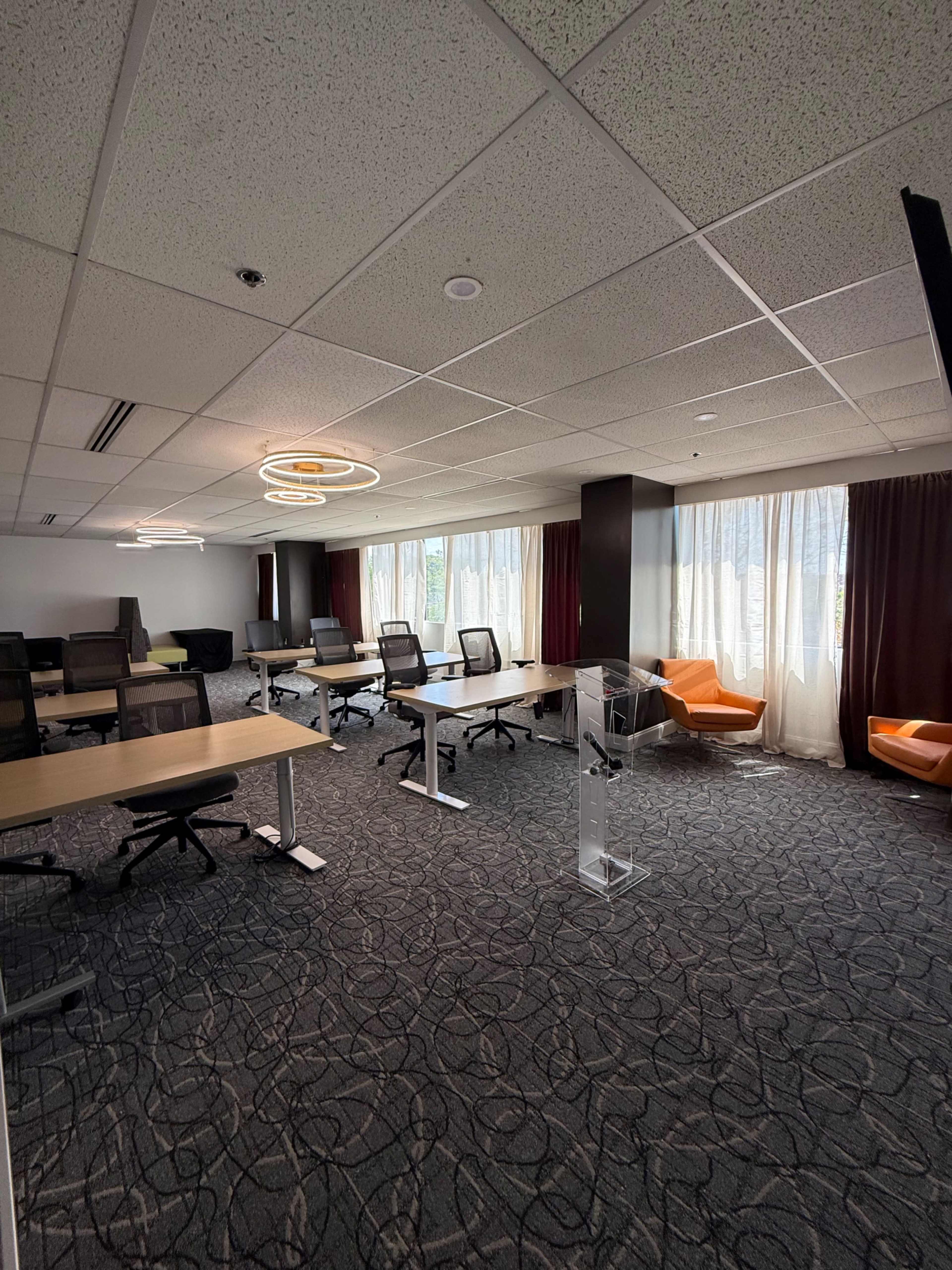The image shows a modern office space with multiple workstations, chairs, and large windows letting in natural light.