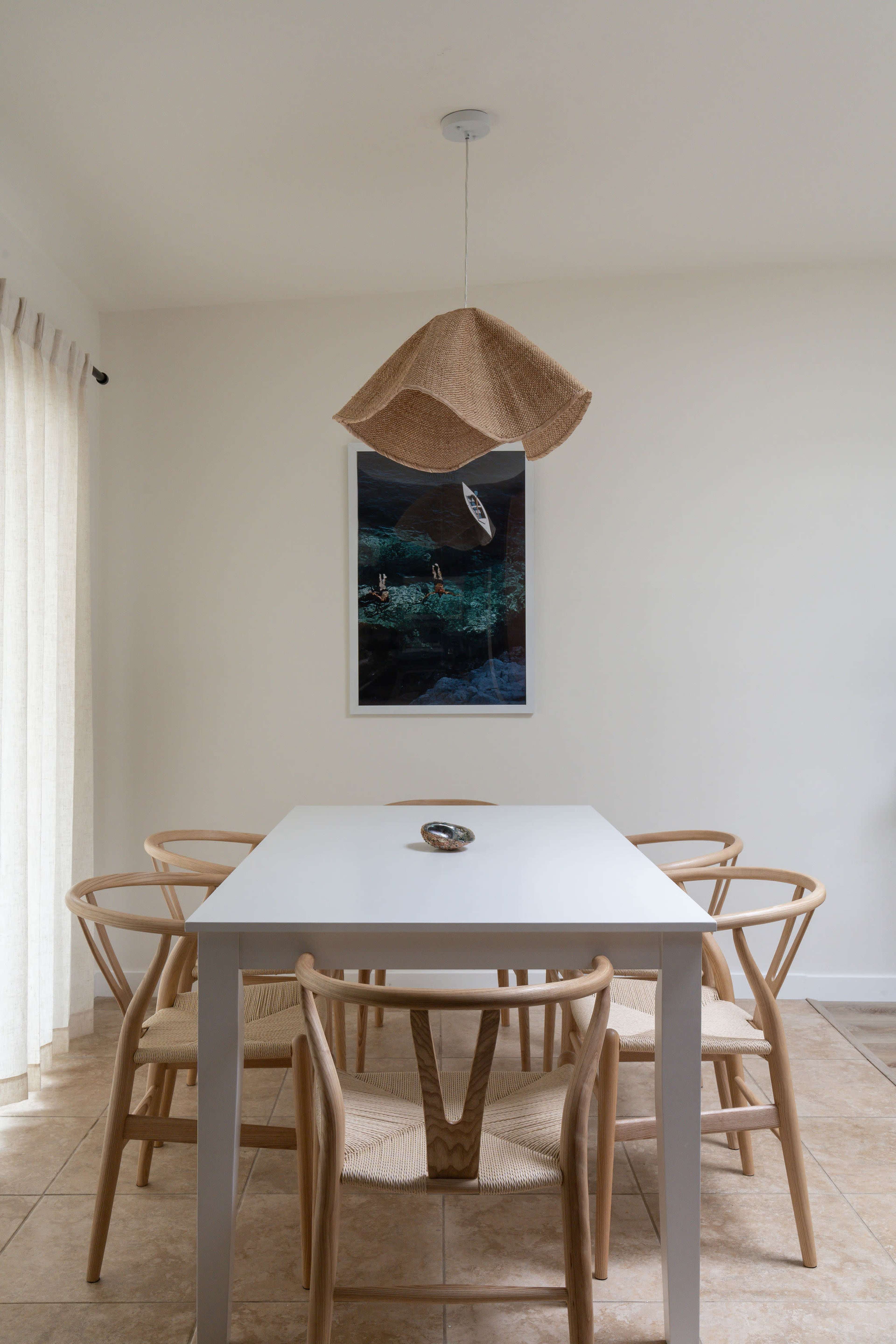 A minimalistic dining area features a white table surrounded by six woven chairs, with a woven pendant light overhead and a framed artwork on the wall.