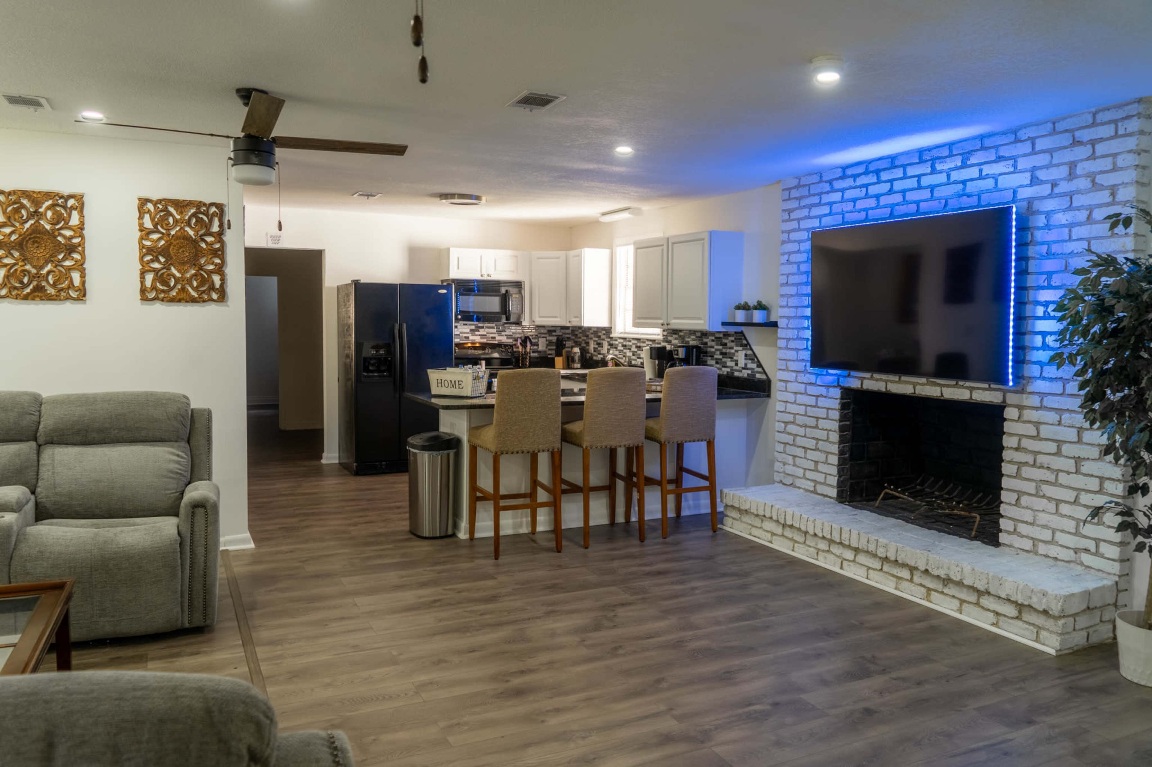 A modern living area with a kitchen and a seating area, featuring a fireplace and a large television mounted on a white brick wall.