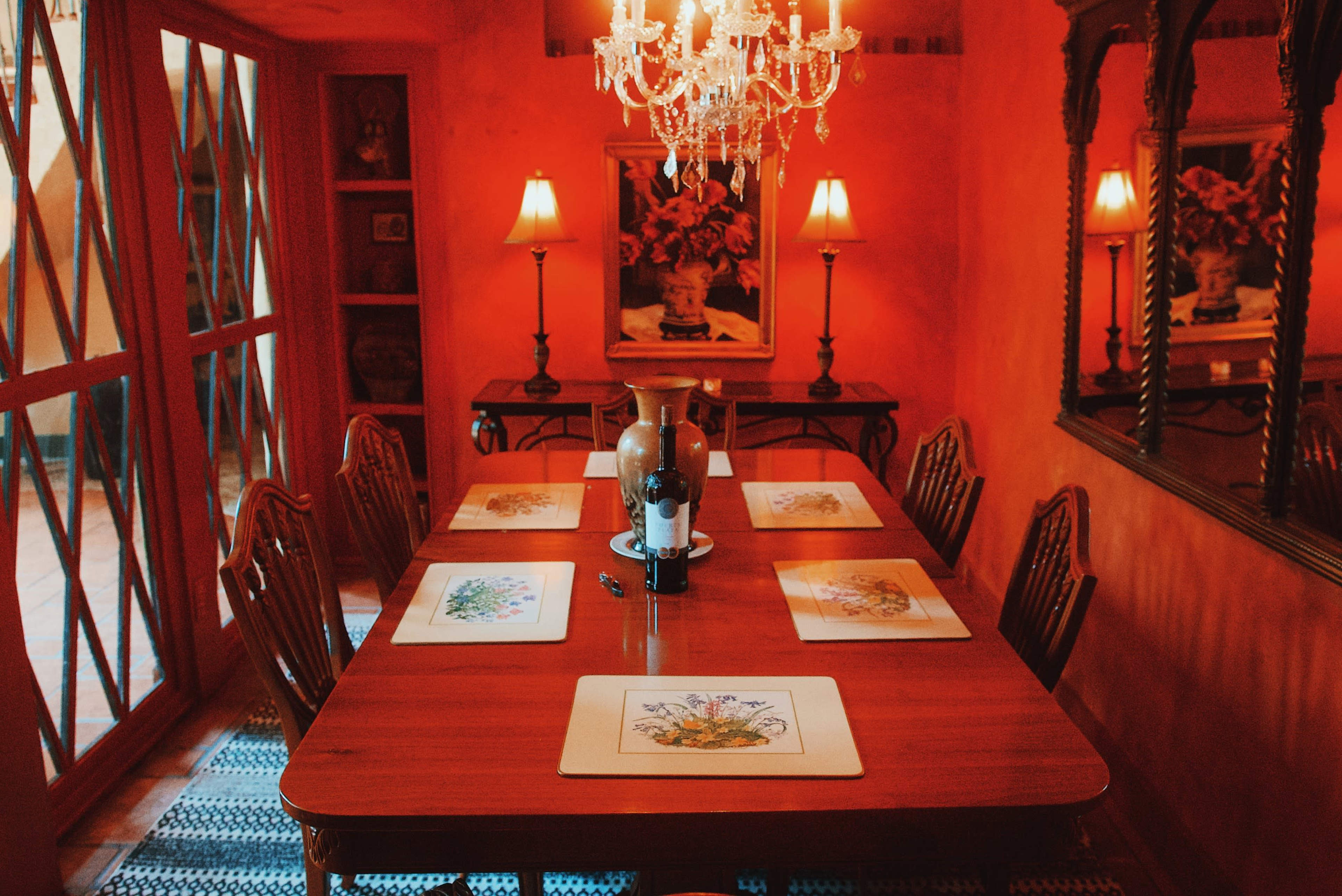 A wooden dining table with eight place settings is illuminated by a chandelier in a warm, red-walled dining room.