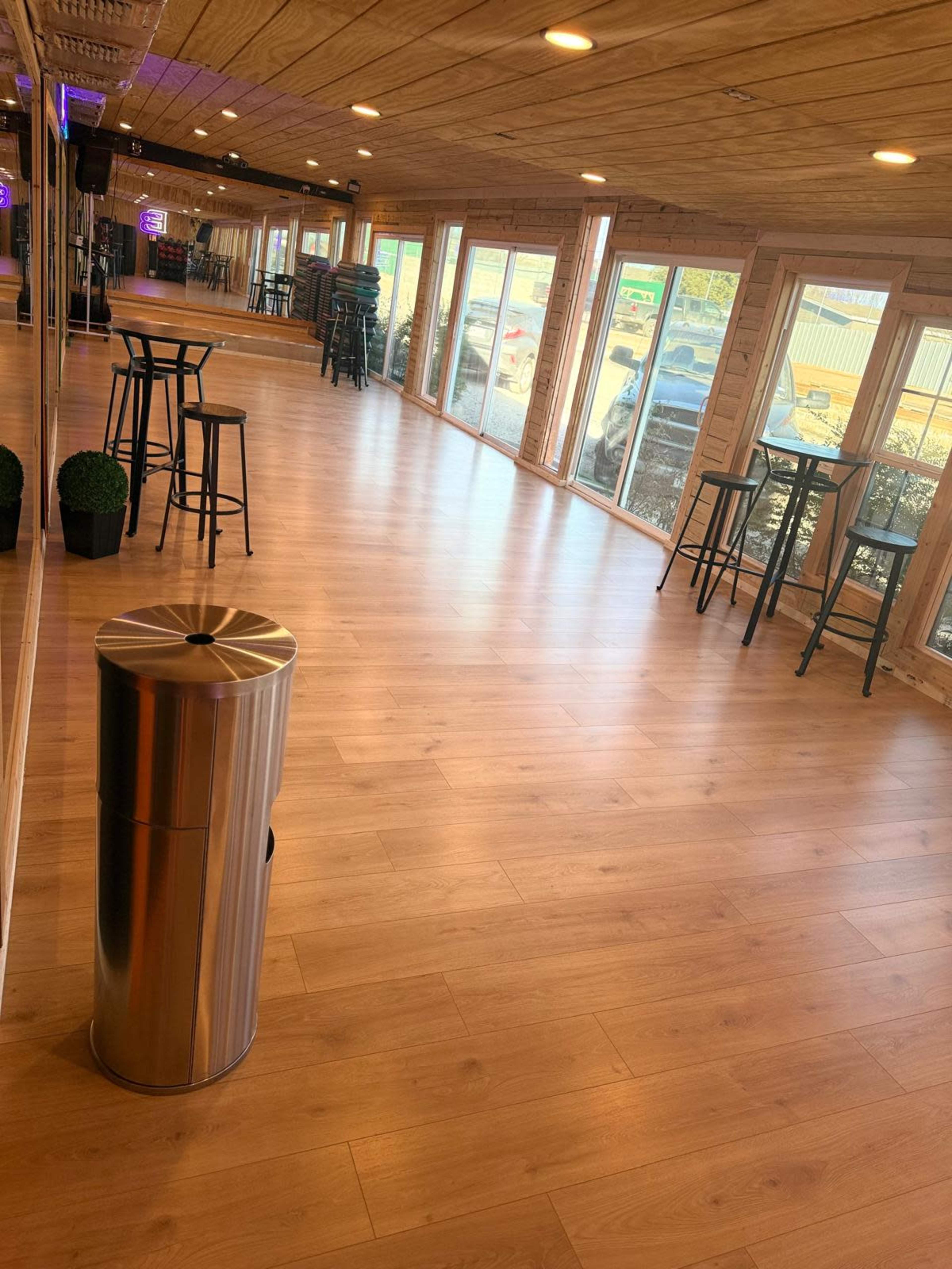 The image shows a spacious interior with wooden flooring, a row of windows, and a few high tables with stools.