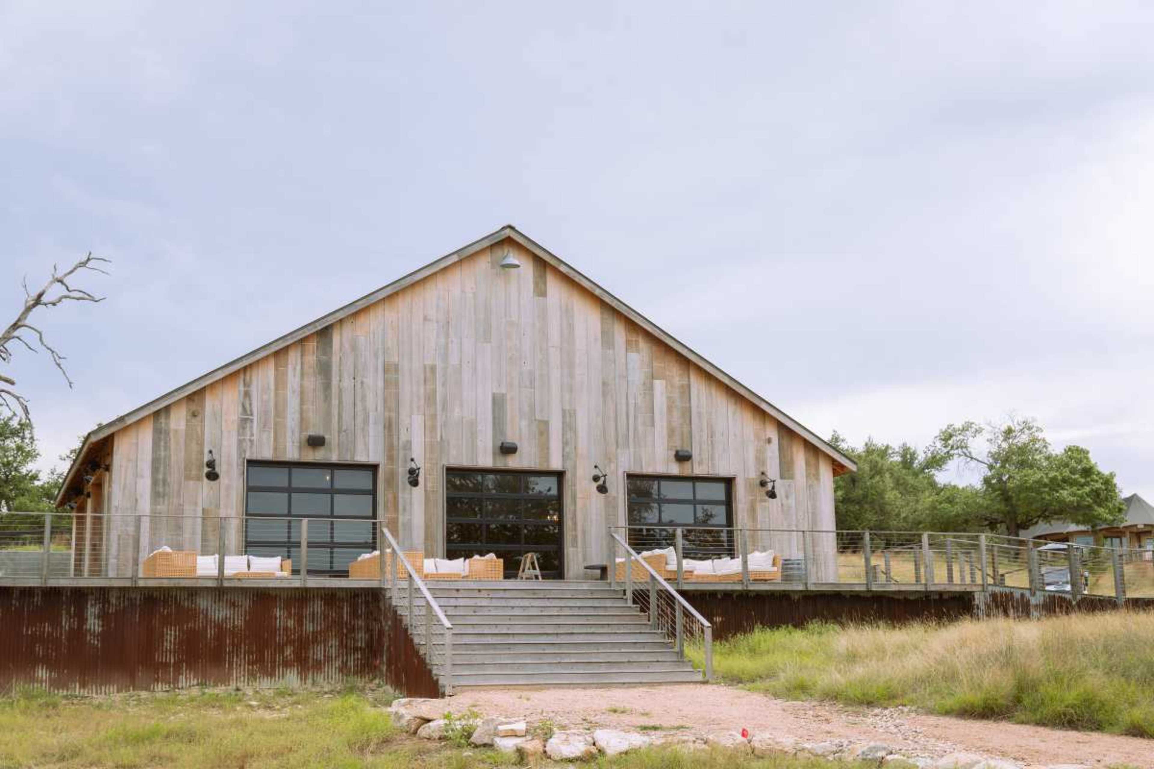 A modern wooden cabin features large windows and a staircase leading up to a deck, surrounded by grass and trees.