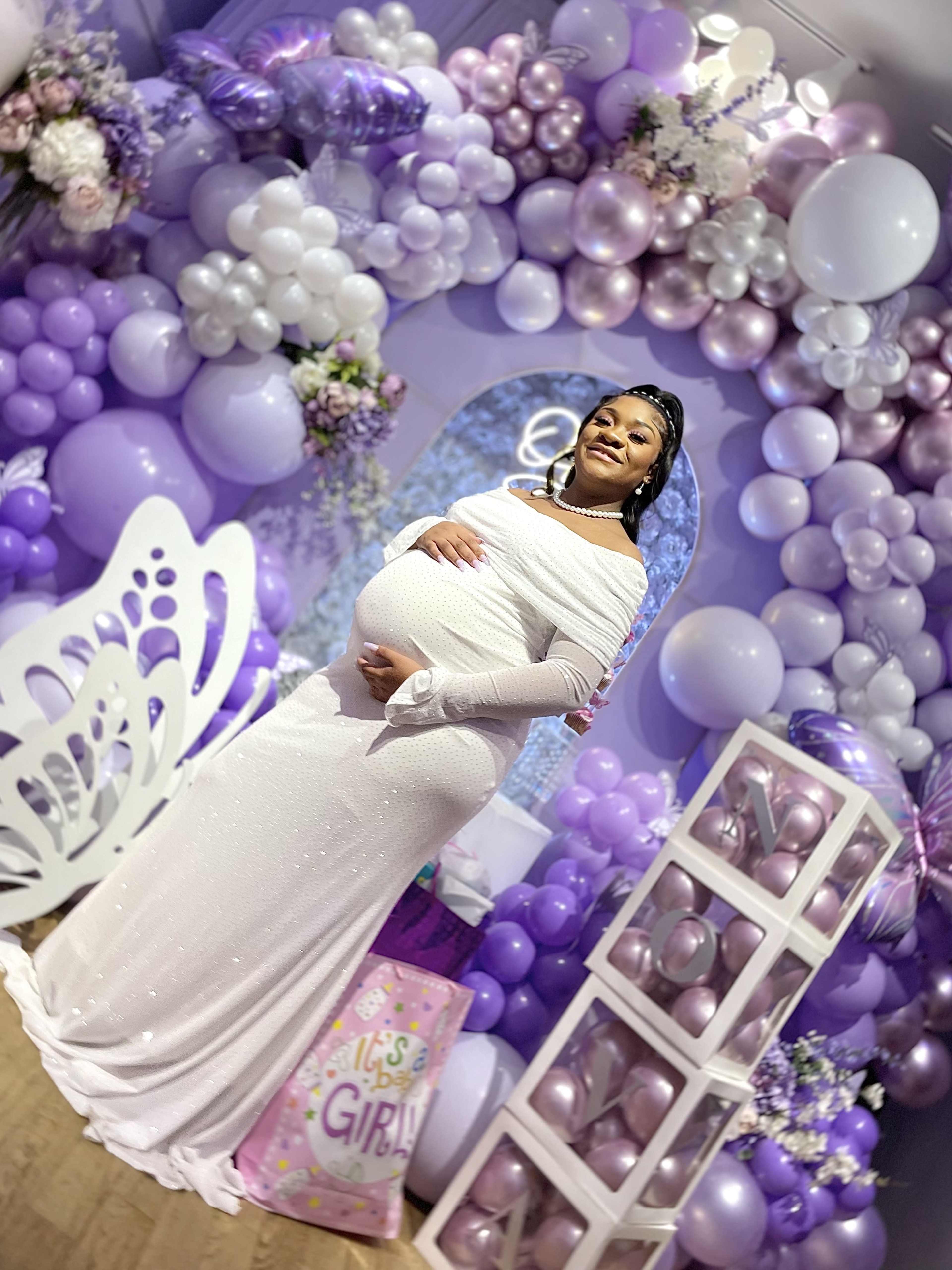 A pregnant woman in a white dress stands in front of a decorative backdrop filled with purple balloons and floral arrangements.