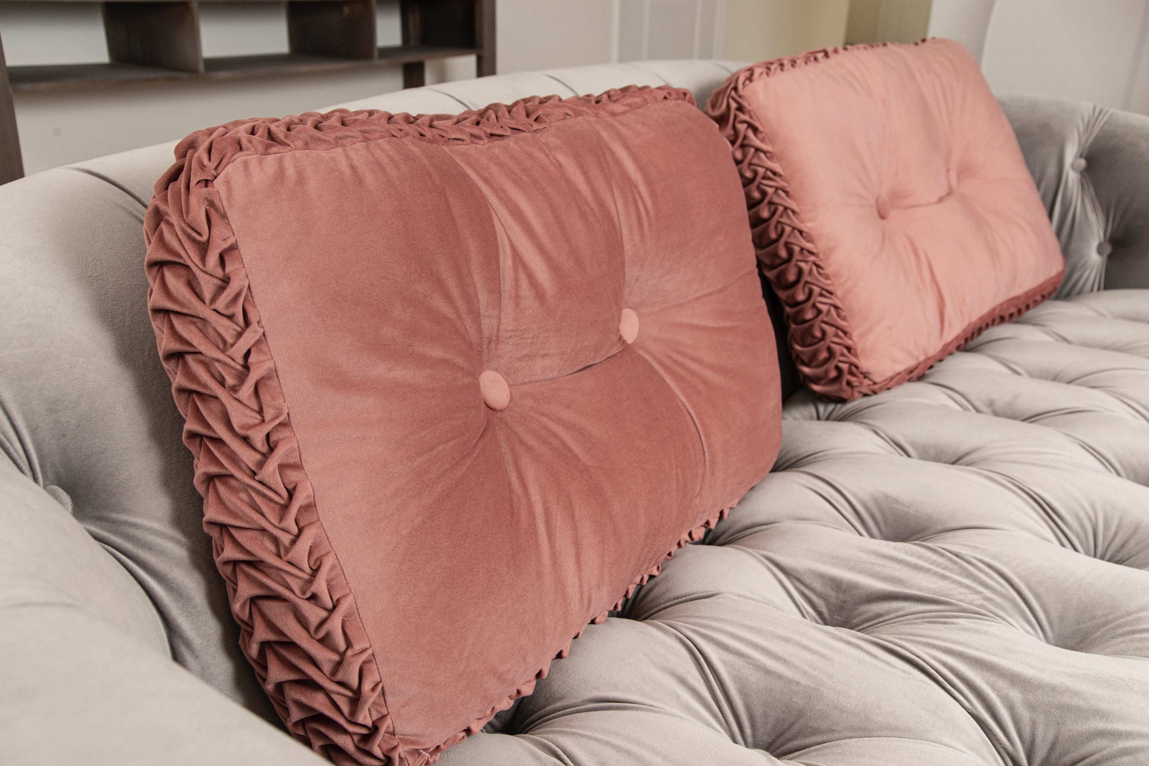 A close-up of a tufted gray sofa with two pink decorative pillows featuring prominent buttons and ruffled edges.