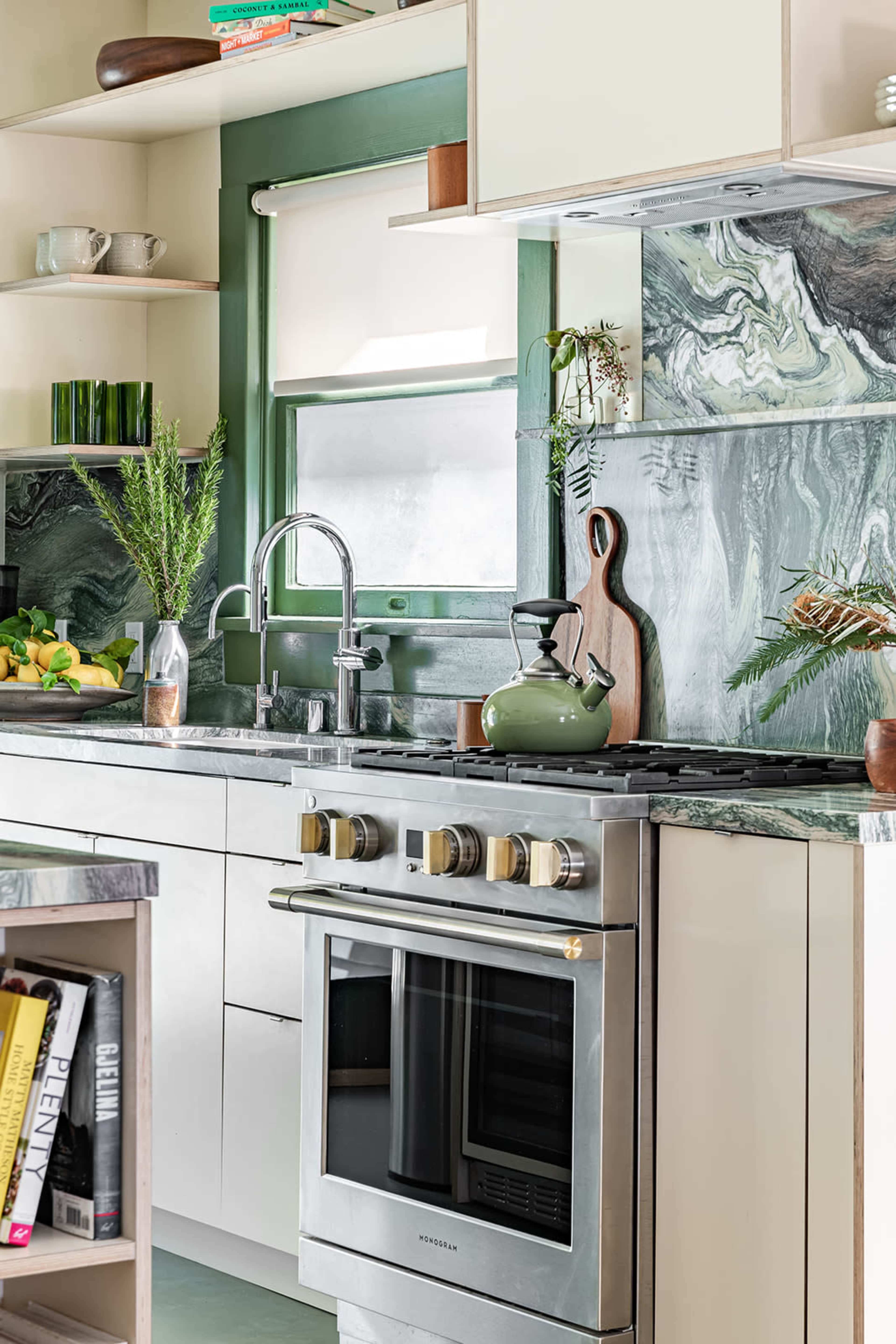 A modern kitchen features green marble countertops, stainless steel appliances, and a window with a view, surrounded by plants and fruit.