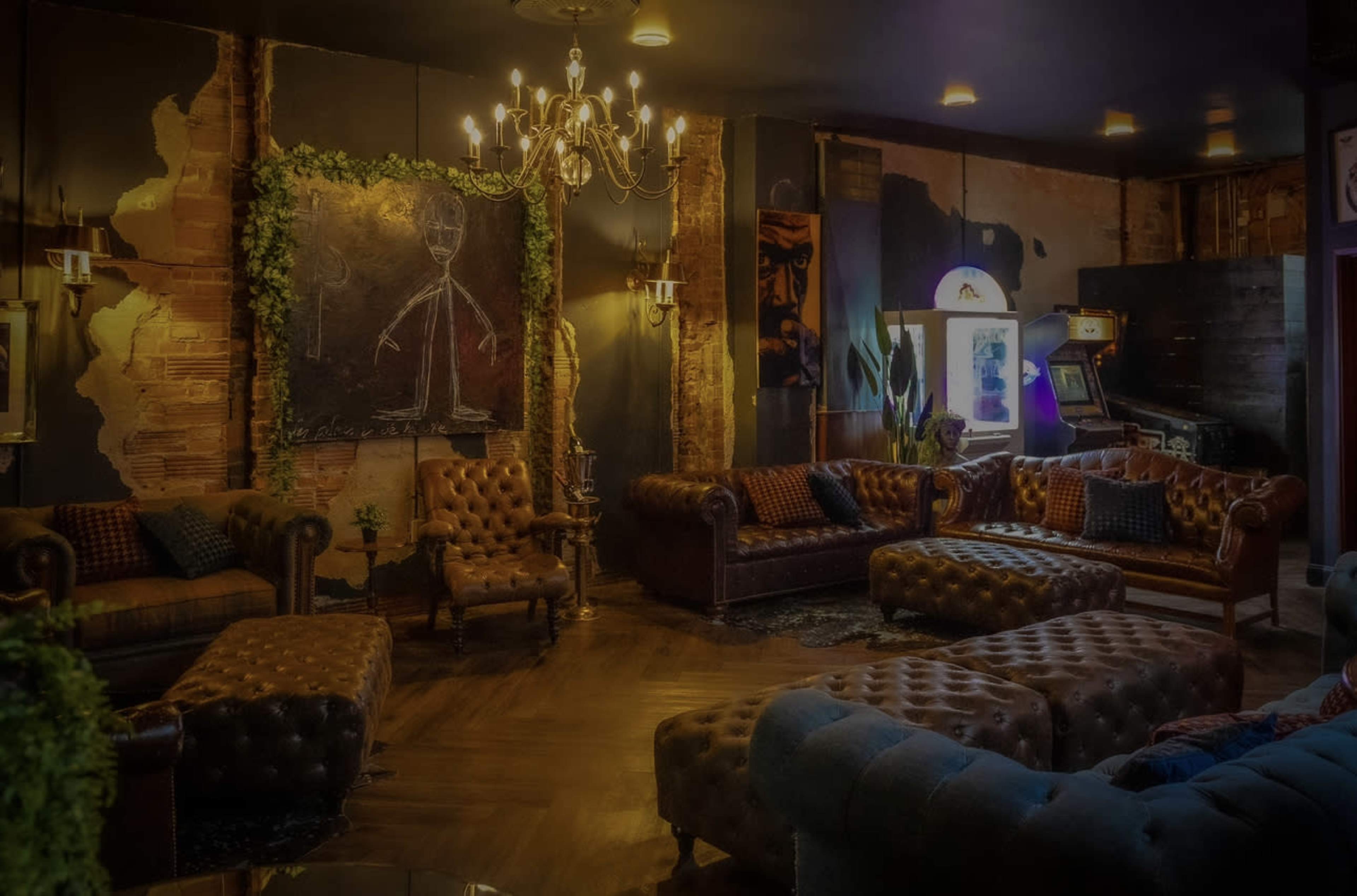 A dimly lit lounge area with leather sofas, a chandelier, and an arcade machine in a rustic setting.