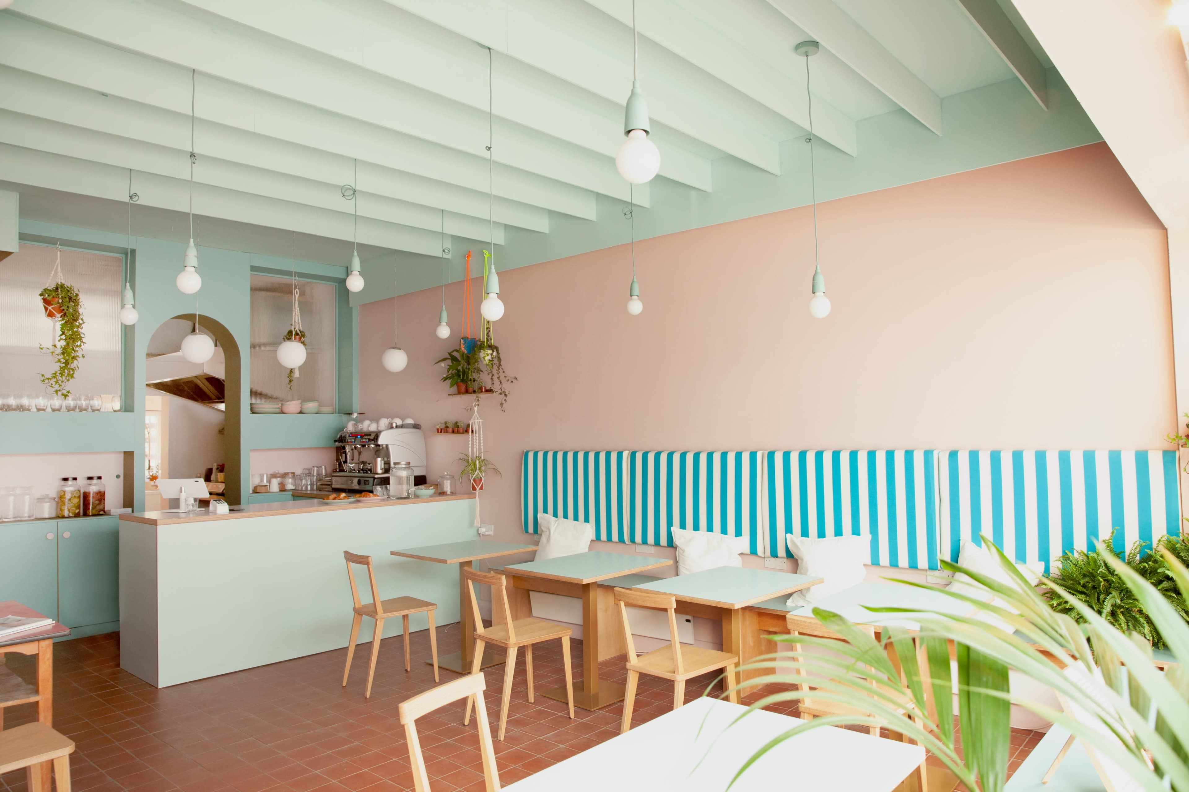 A modern cafe features light blue and pink walls, wooden tables, and striped seating with hanging pendant lights.