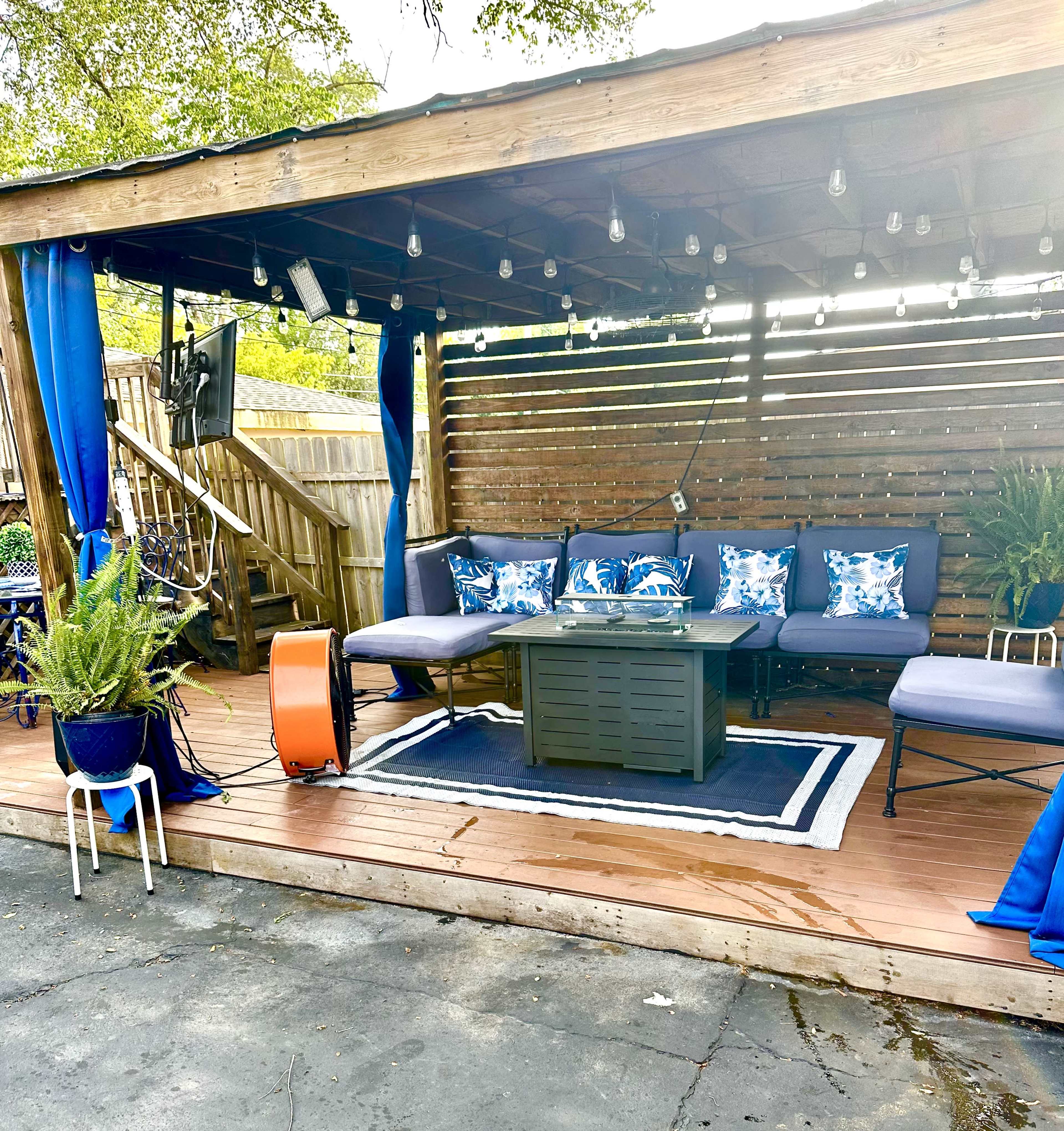 A wooden patio area with a sofa, a coffee table, and decorative pillows, surrounded by string lights and potted plants.