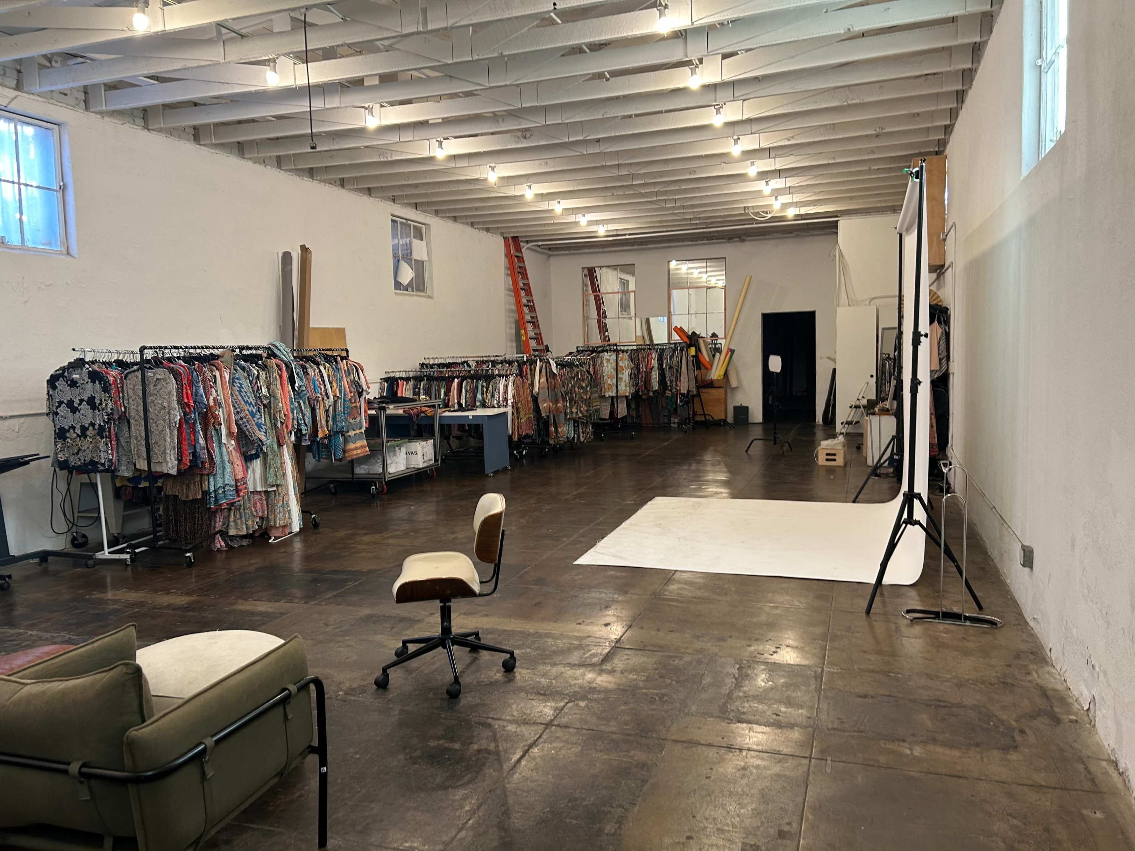 A spacious studio features racks of clothing along one wall, a white backdrop on the floor, and a chair in the foreground.
