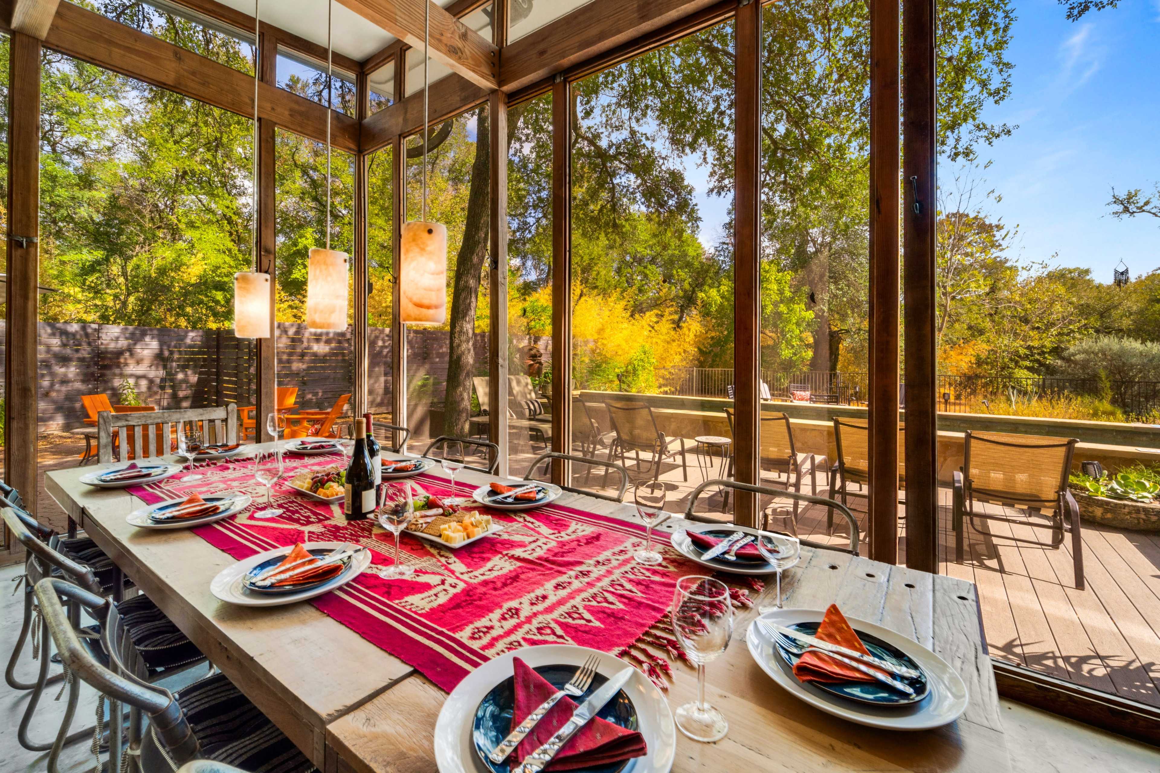 A dining table is set with plates and glasses in a glass-enclosed room, overlooking a patio and lush greenery outside.