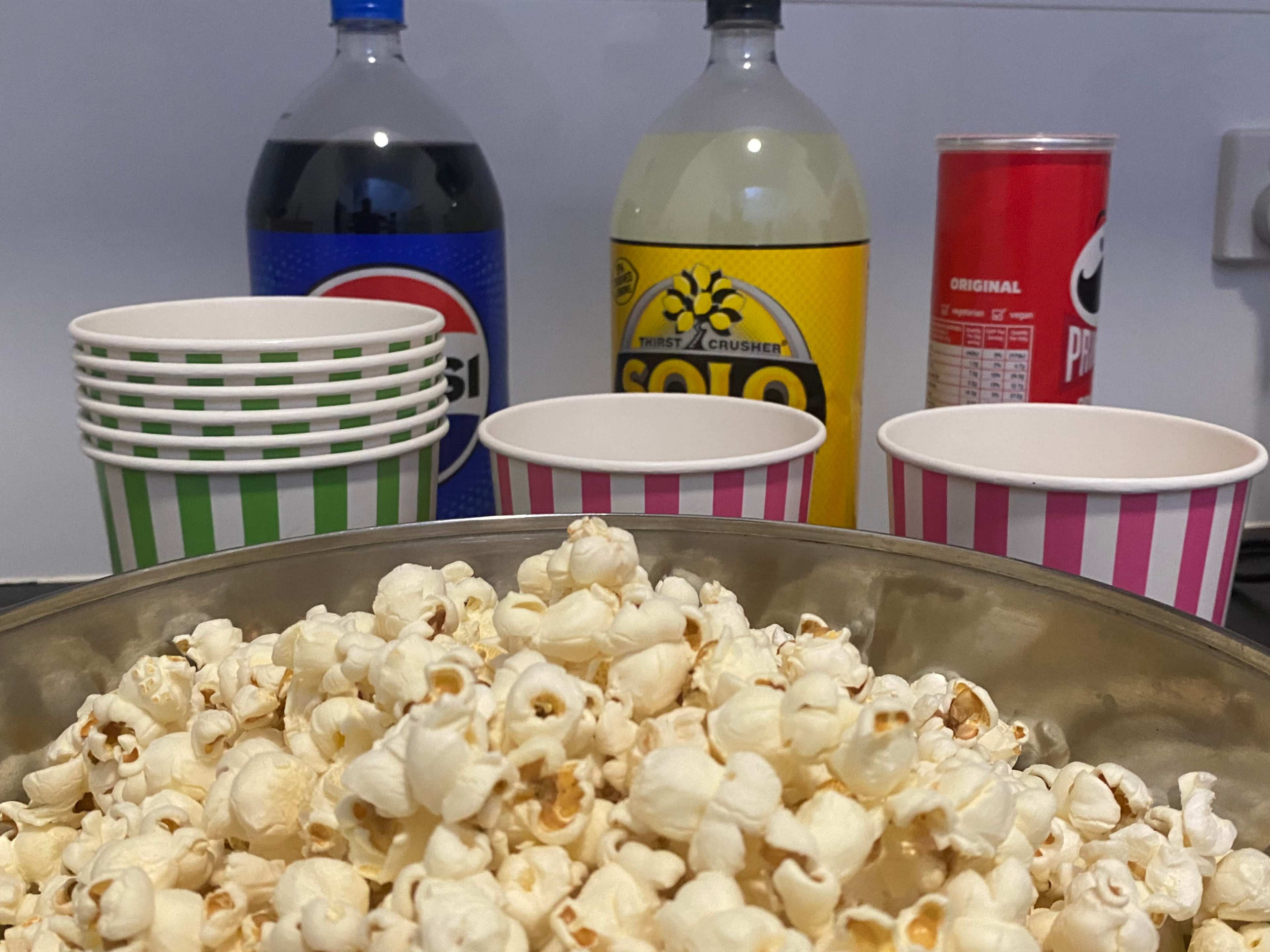 The image shows a large bowl of popcorn in the foreground, with colorful striped cups and bottles of soda in the background.