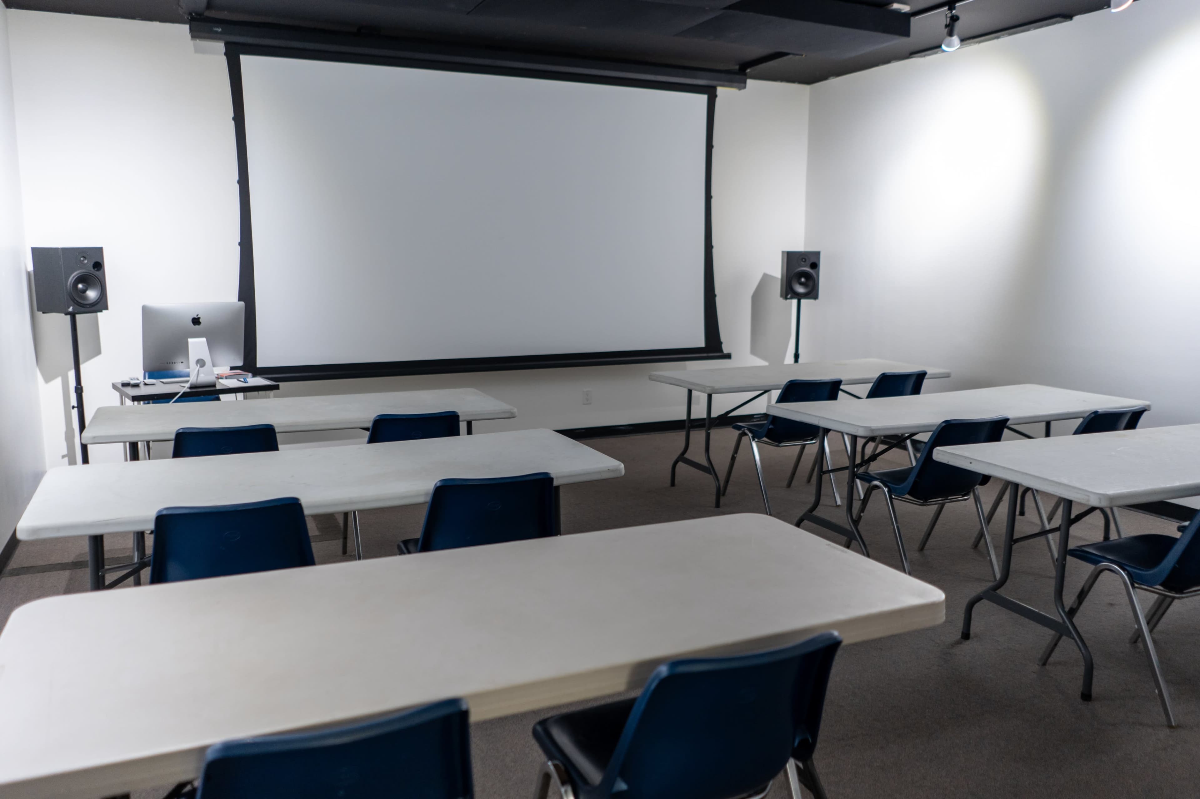 The image shows a classroom setup with several rectangular tables and chairs facing a large projection screen at the front.