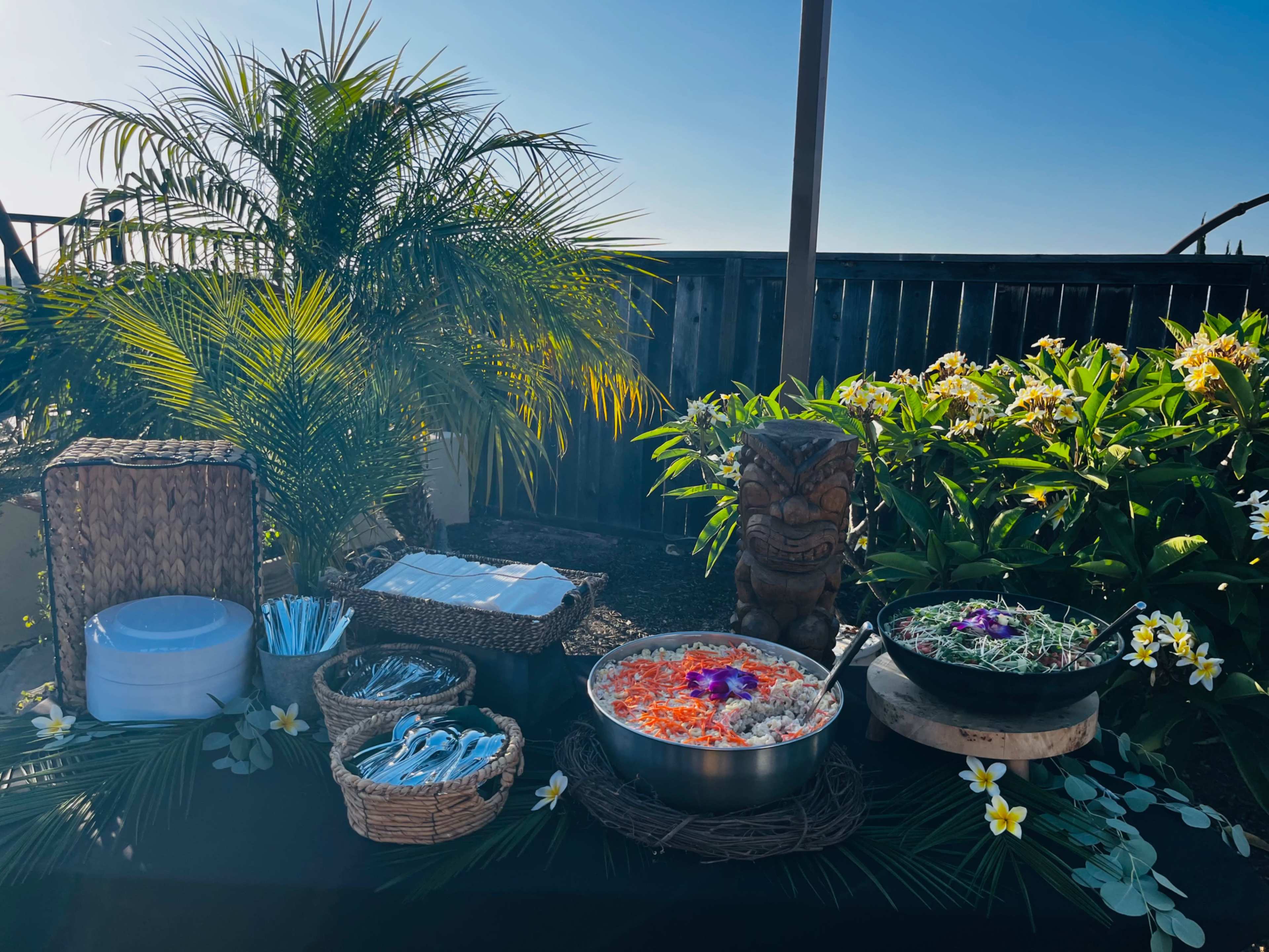 A buffet table is set up outdoors, featuring bowls of colorful food surrounded by tropical plants and decorations.