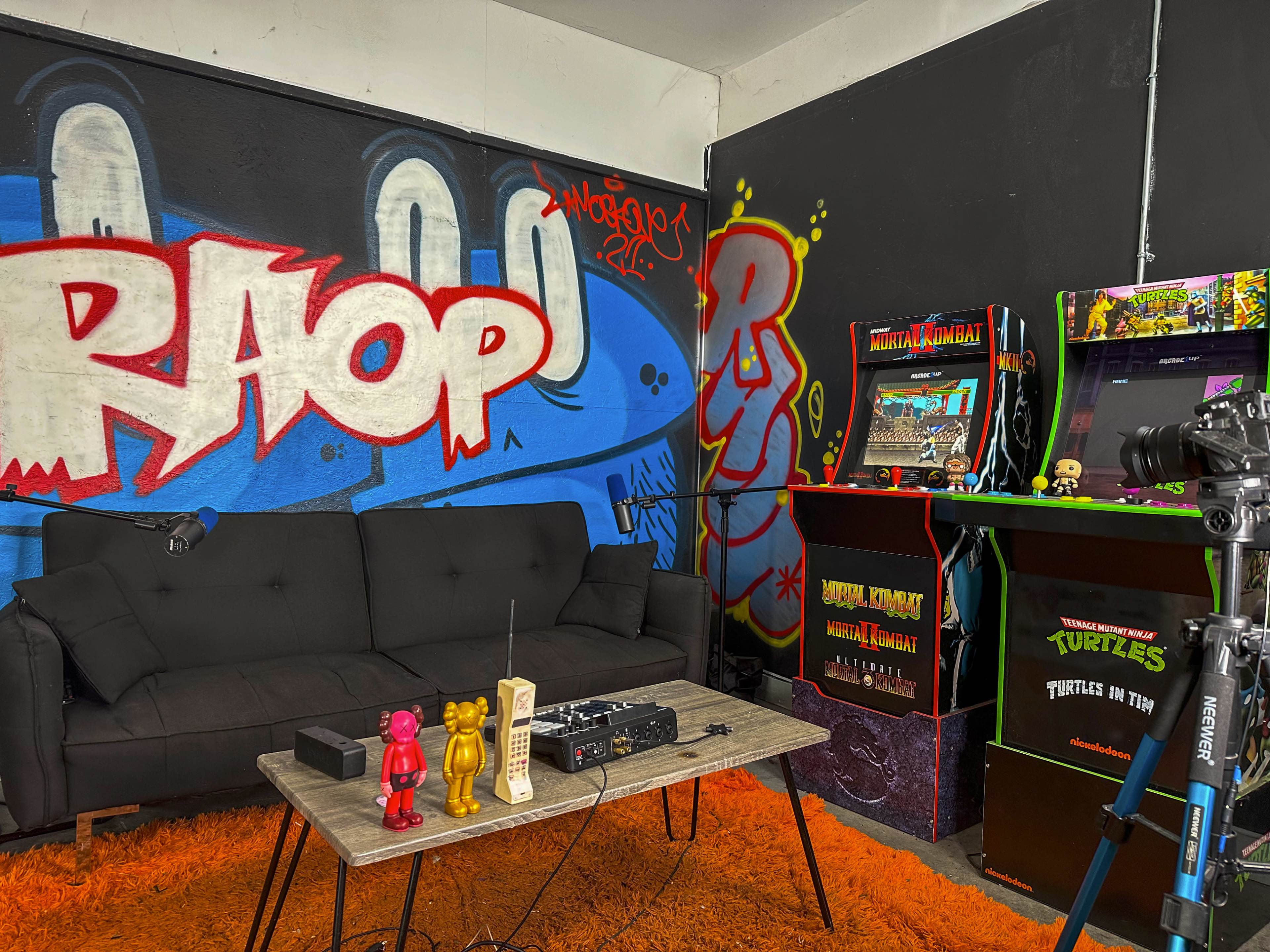 The scene features a lounge area with a black couch, a coffee table adorned with colorful toys and an old telephone, and arcade machines against a graffiti-covered wall.