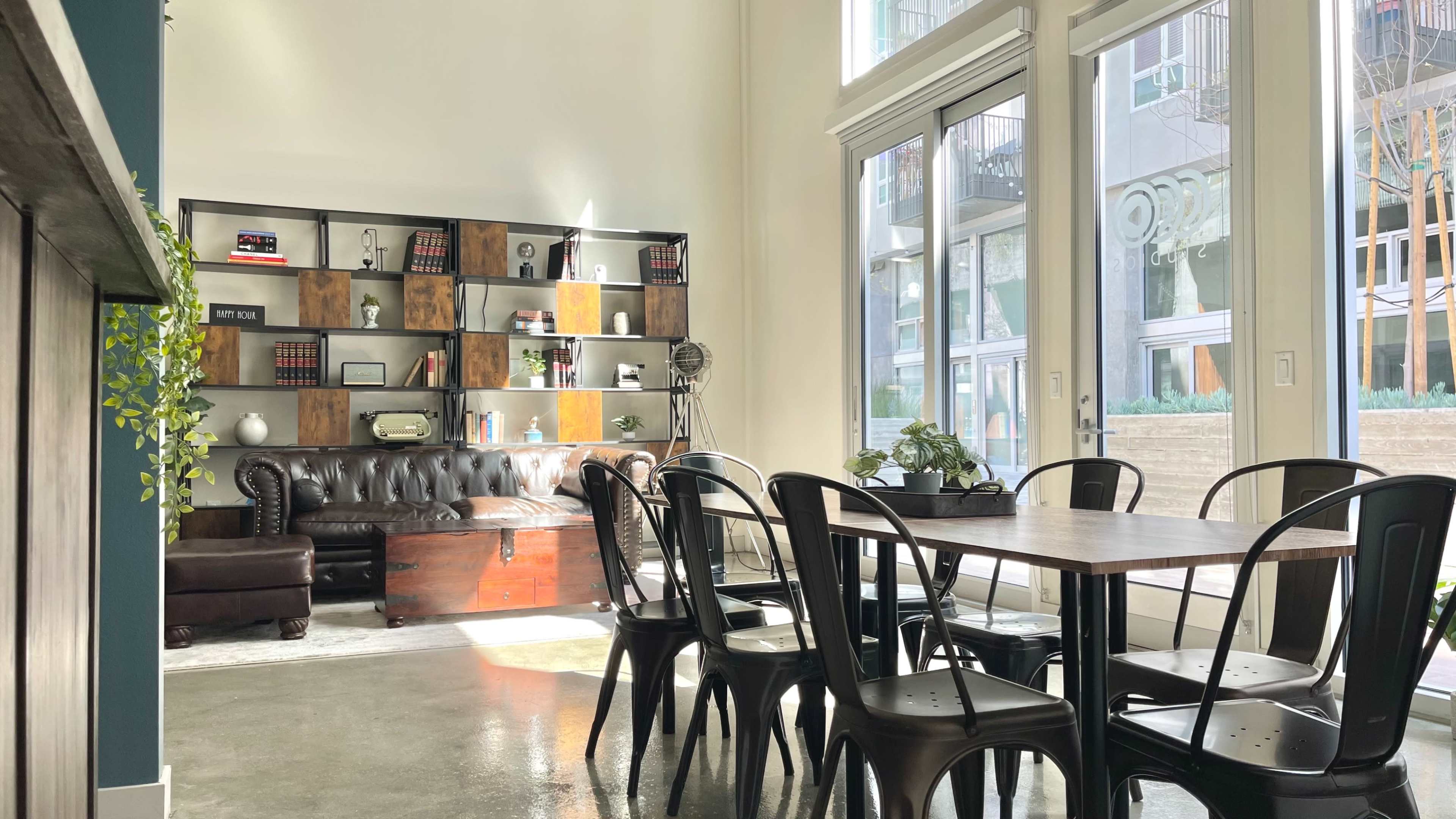 A modern interior space featuring a leather couch, a wooden coffee table, a bookshelf with various items, and a row of metal dining chairs surrounding a wooden table.